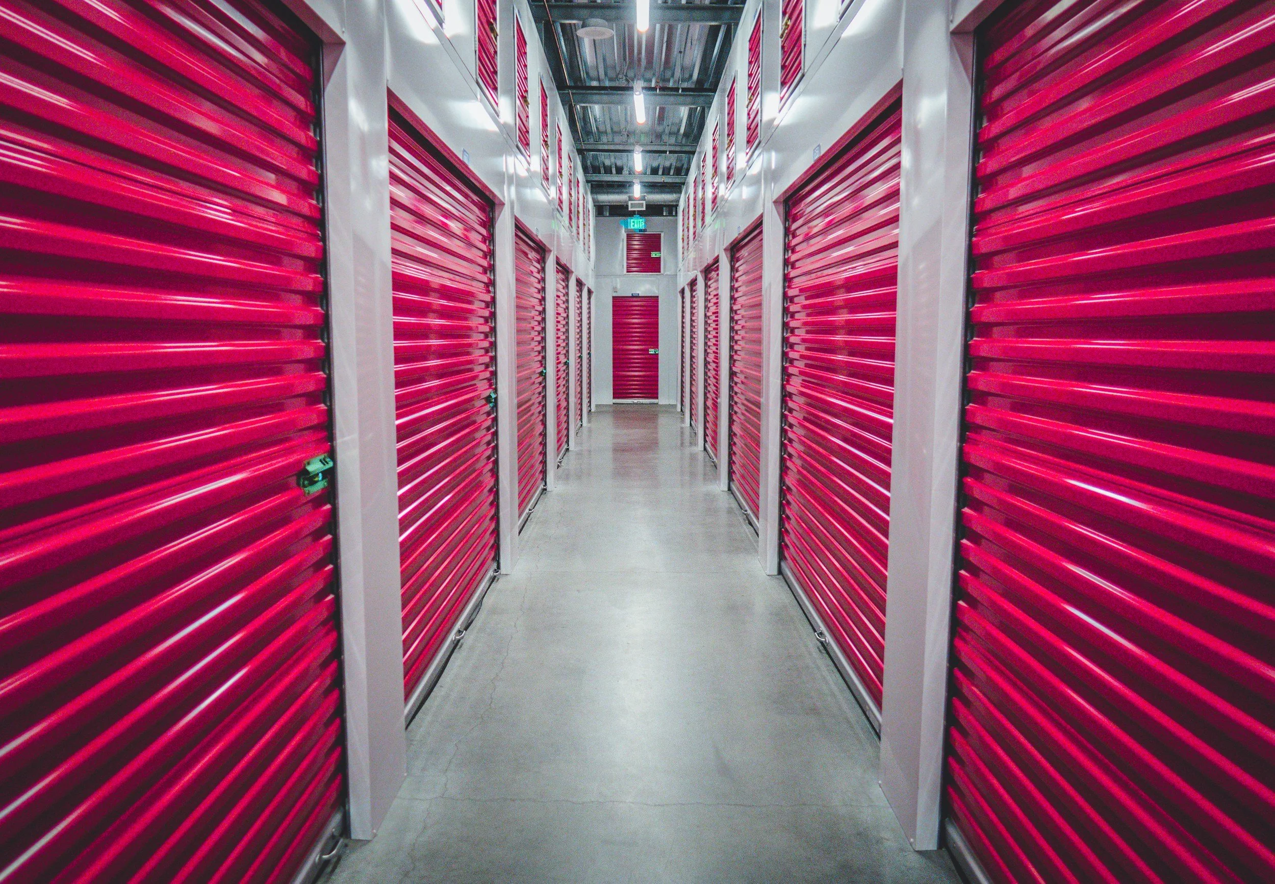 A hallway inside a storage facility with red roll-up doors on each side. The hallway has a concrete floor, white walls, and a ceiling with exposed industrial piping and lighting. An exit sign is visible at the end of the hallway.