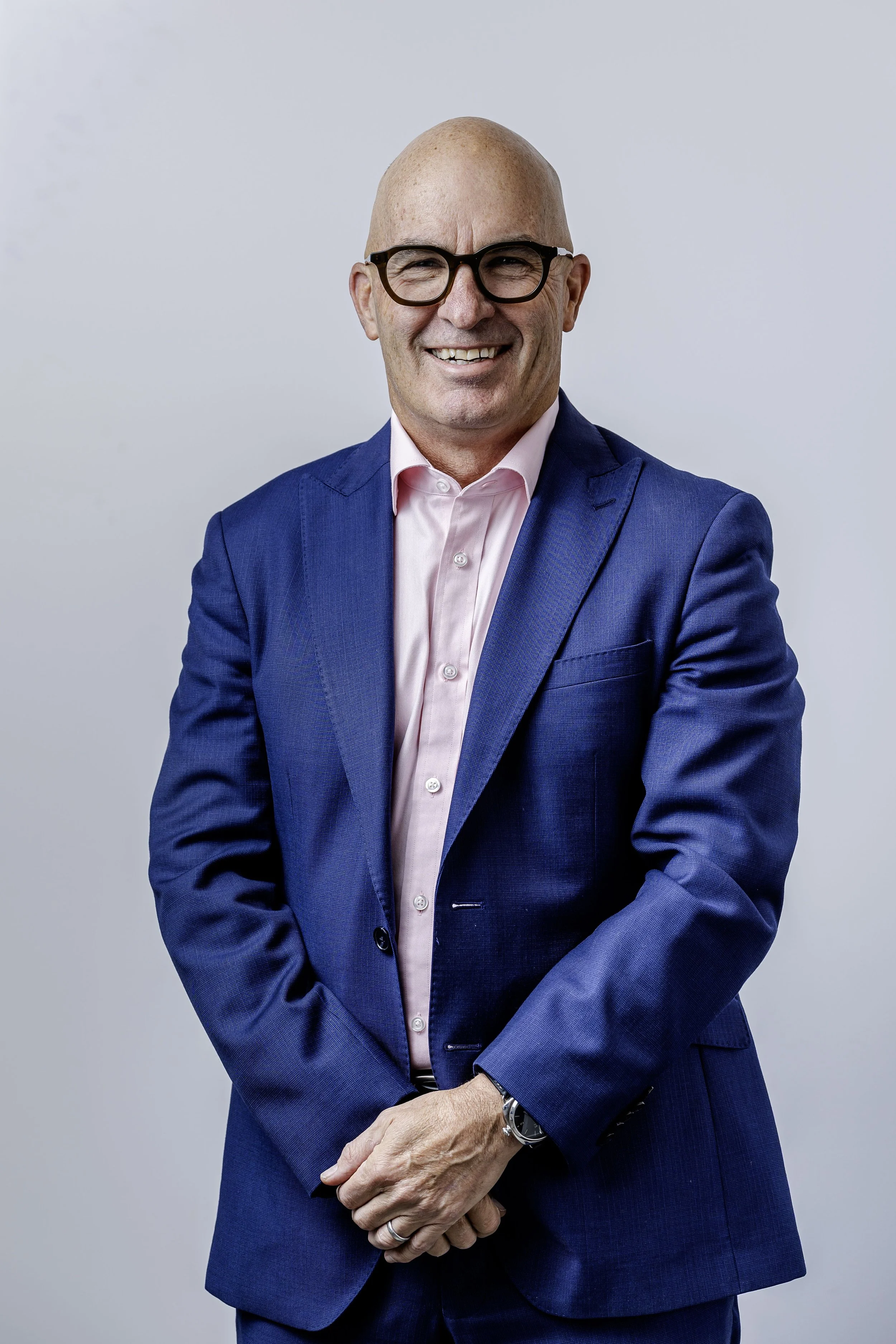 A middle-aged man with a bald head and glasses, wearing a blue suit and pink shirt, smiling and posing with his arms crossed against a plain light background.