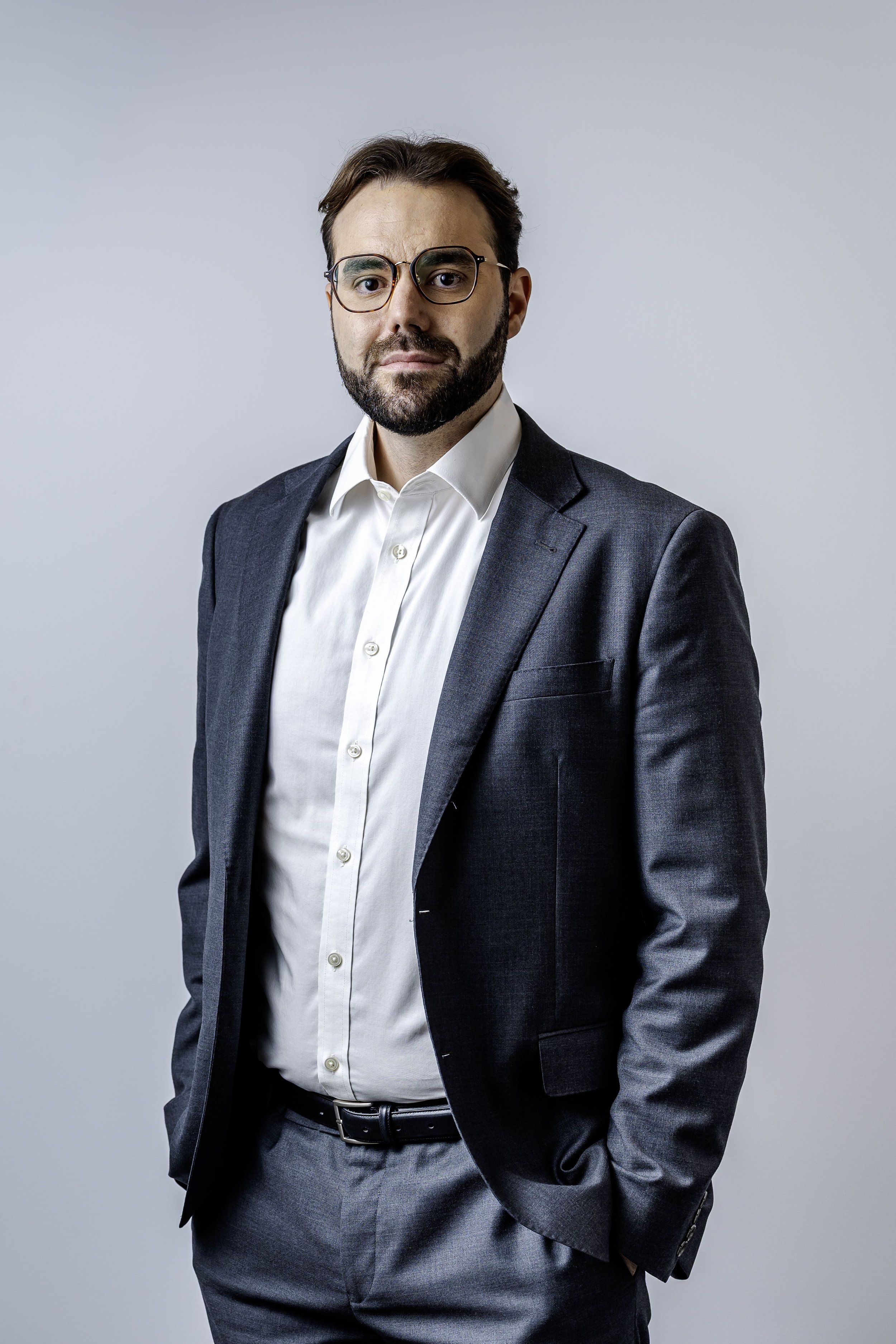 A man with dark hair, beard, wearing glasses, a grey suit, and a white shirt, standing against a plain light grey background.
