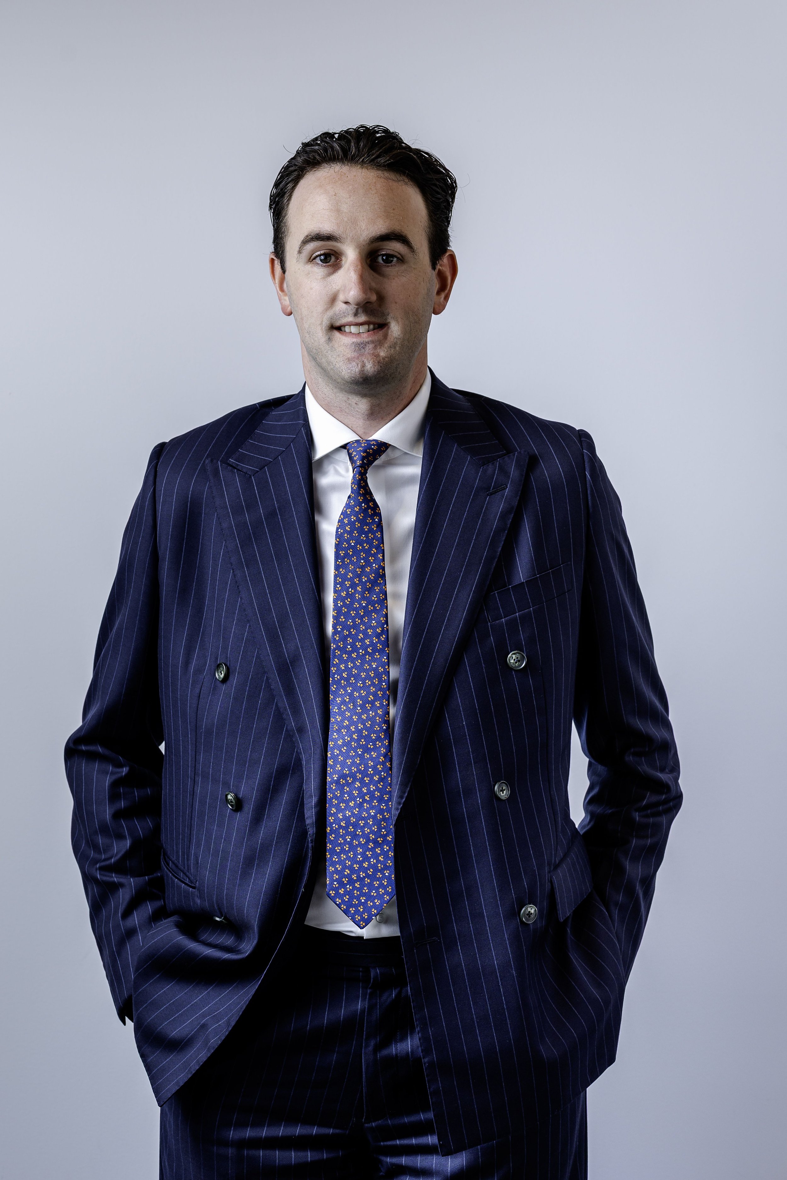 A young man with dark hair dressed in a navy blue pinstripe suit, white shirt, and a blue tie with small orange and yellow floral patterns, standing against a plain light gray background.