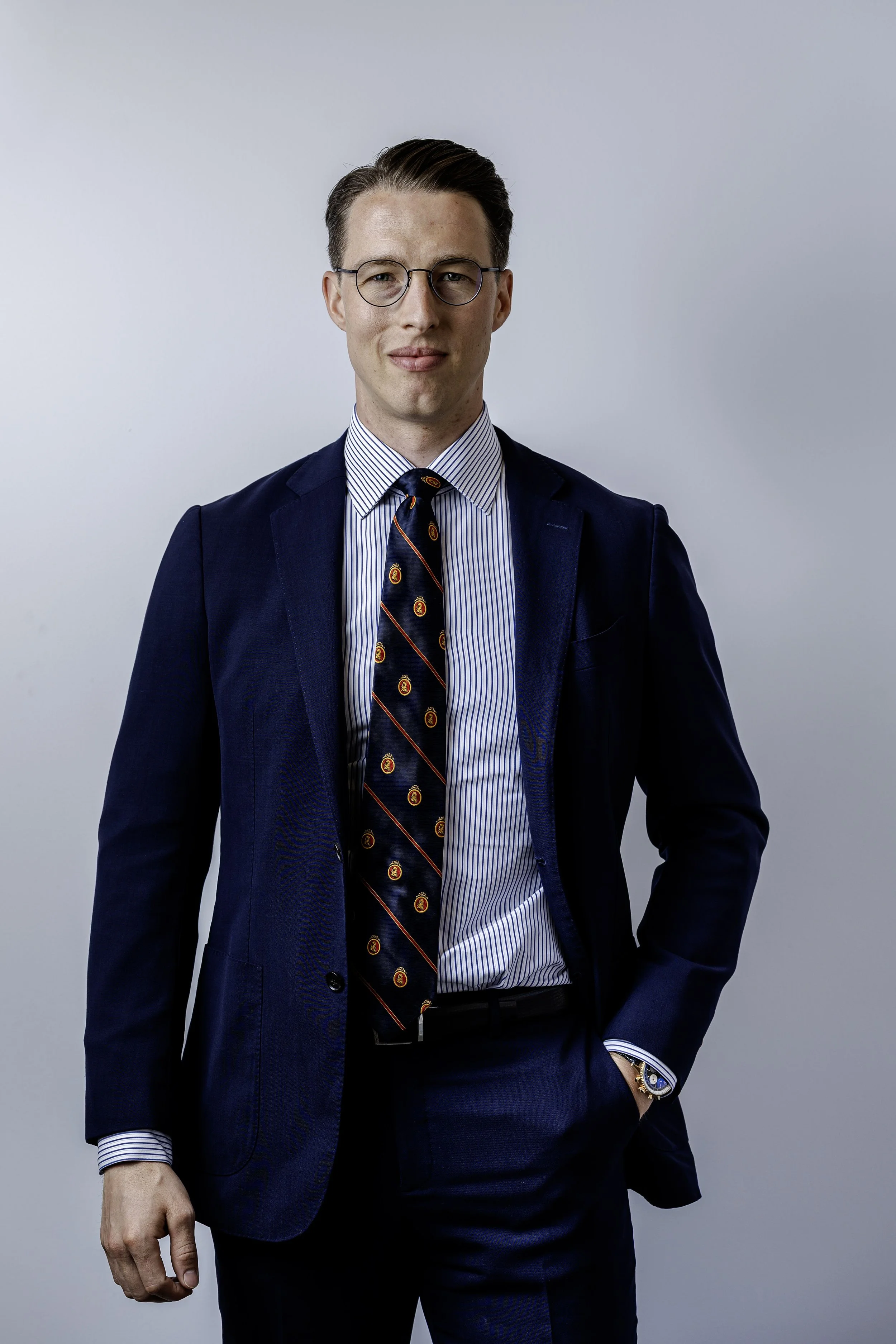 A man wearing glasses, a navy suit, a blue and white striped shirt, and a navy tie with crest designs, standing against a plain light gray background.