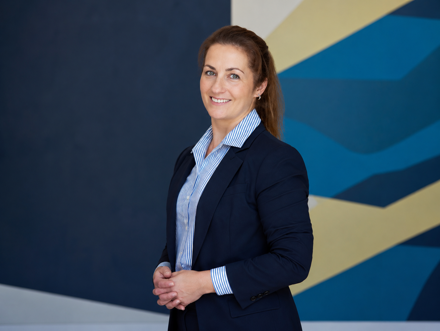 A professional woman in a dark blazer and light blue striped shirt standing in front of a colorful geometric wall. She has brown hair, blue eyes, and is smiling.