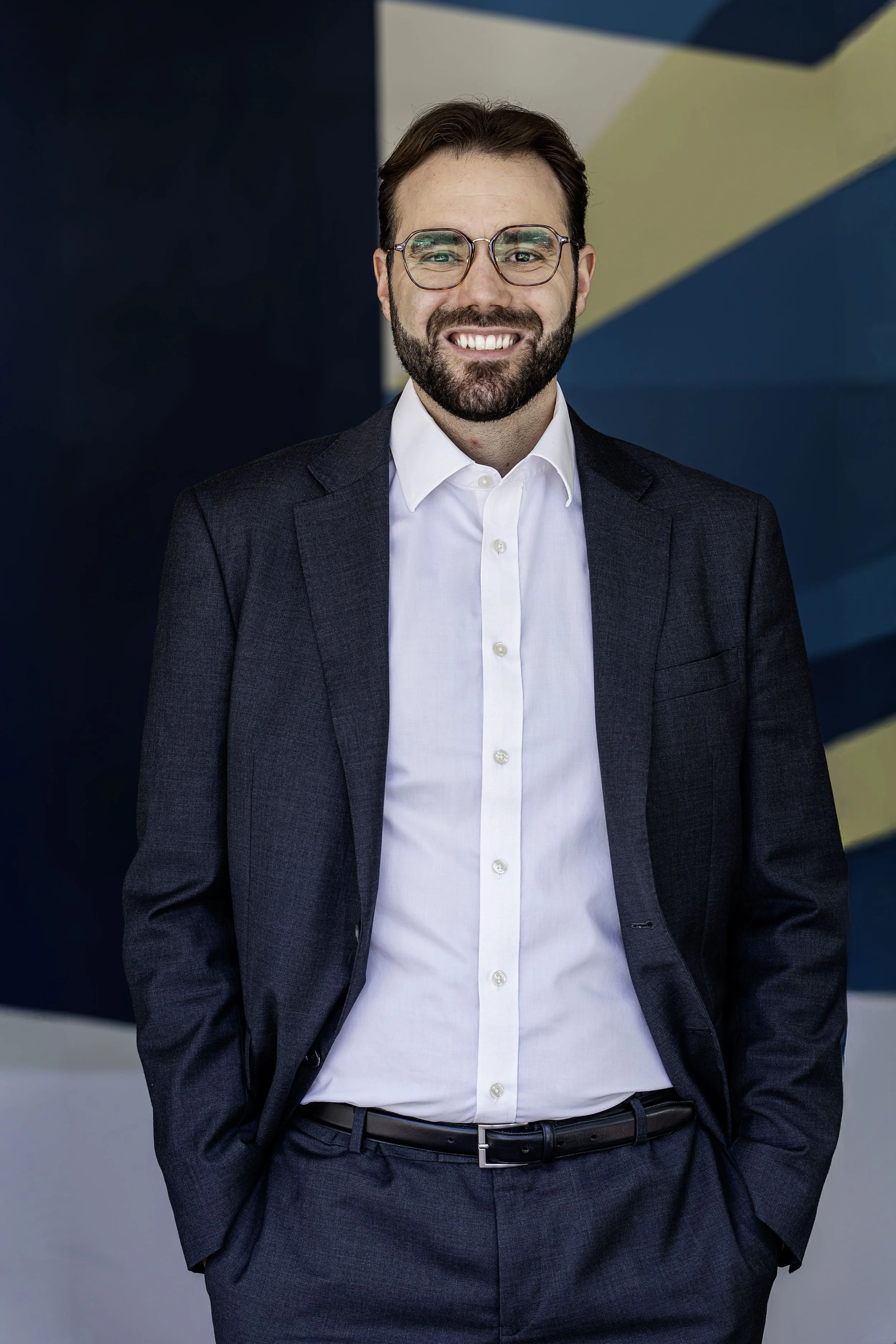A man in a dark gray business suit with a white shirt and black belt, smiling and standing with his hands in his pockets against a blue and yellow geometric background.