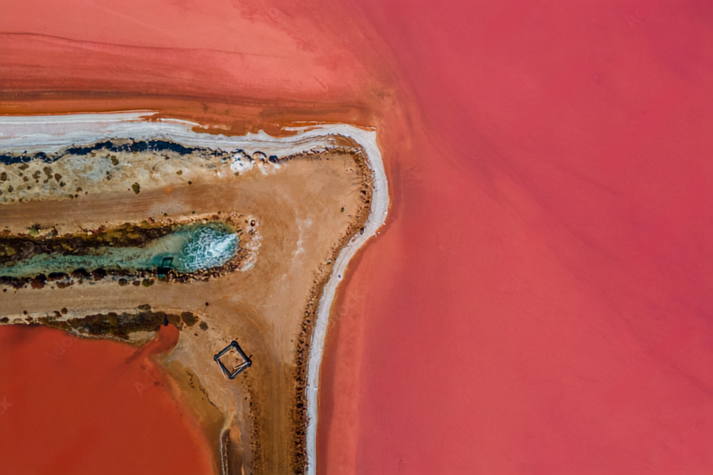 Aerial view of a small natural water body with blue-green water, surrounded by reddish and sandy terrain, with some vegetation and a small square structure on the land nearby.