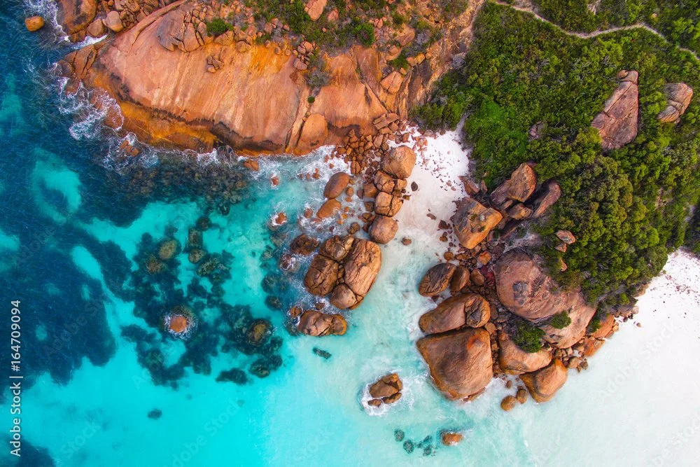 Aerial view of a tropical coastline with clear blue waters, large reddish-brown rocks, and dense green vegetation.