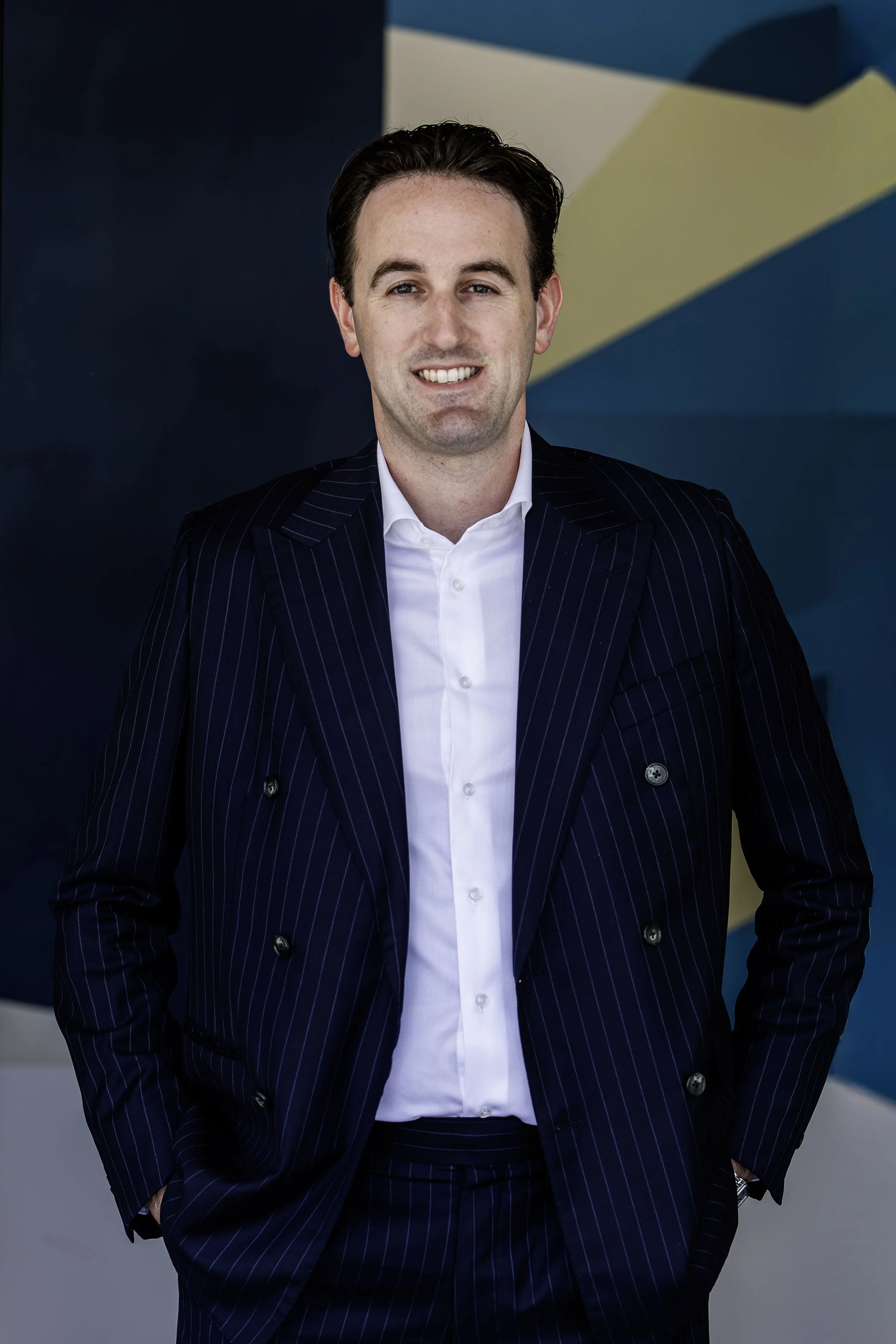 Man in a navy pinstripe suit with white shirt standing against a background with blue and yellow geometric shapes.