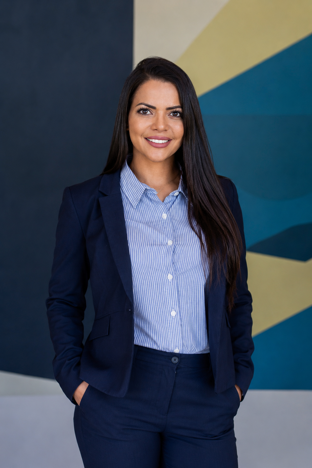A woman with long dark hair wearing a navy blazer and striped blue shirt, smiling and posing with her hands in her pockets against a geometric painted wall.