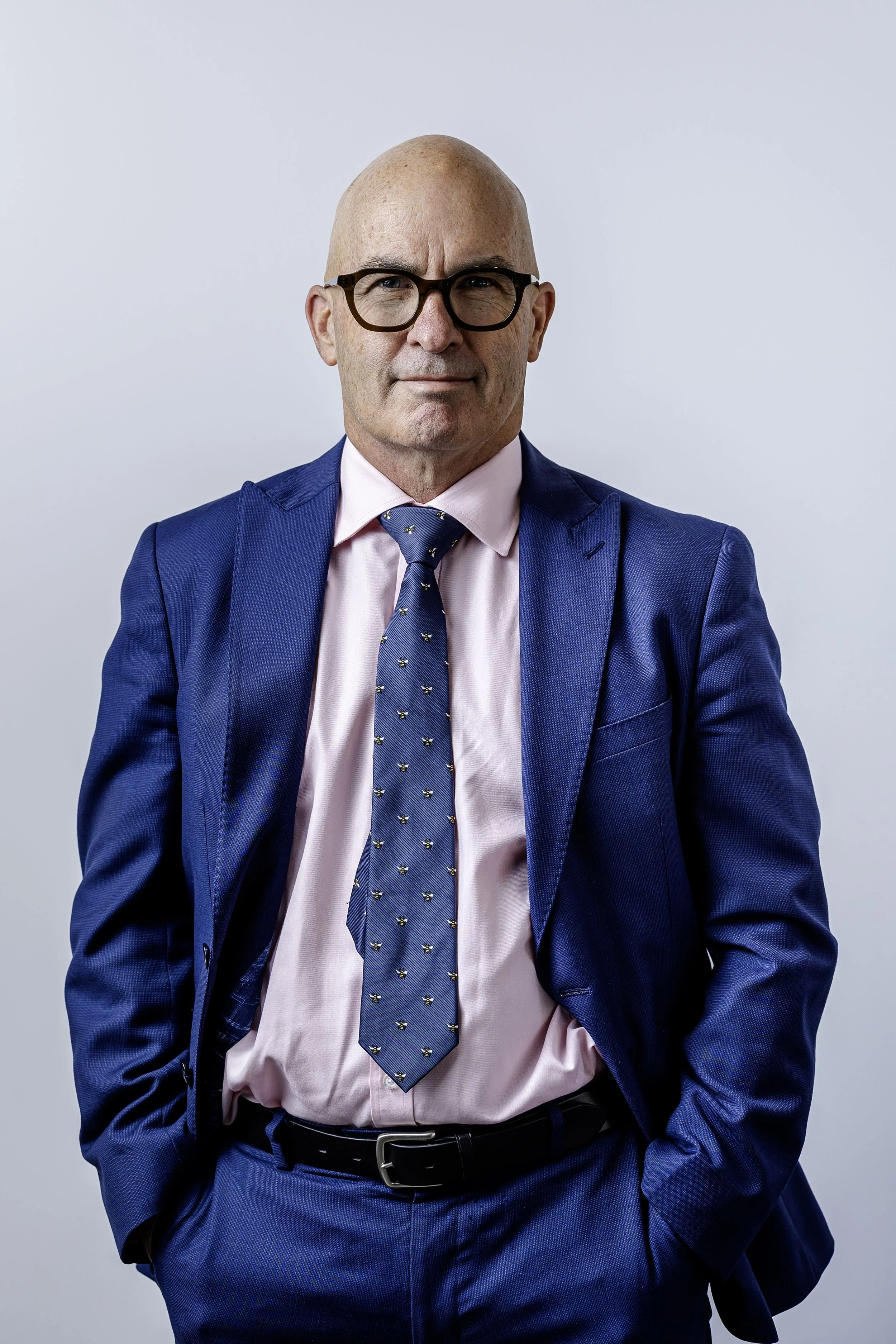 A bald man wearing glasses, a pink dress shirt, a blue suit, and a matching blue tie with small yellow patterns, standing against a plain light gray background.