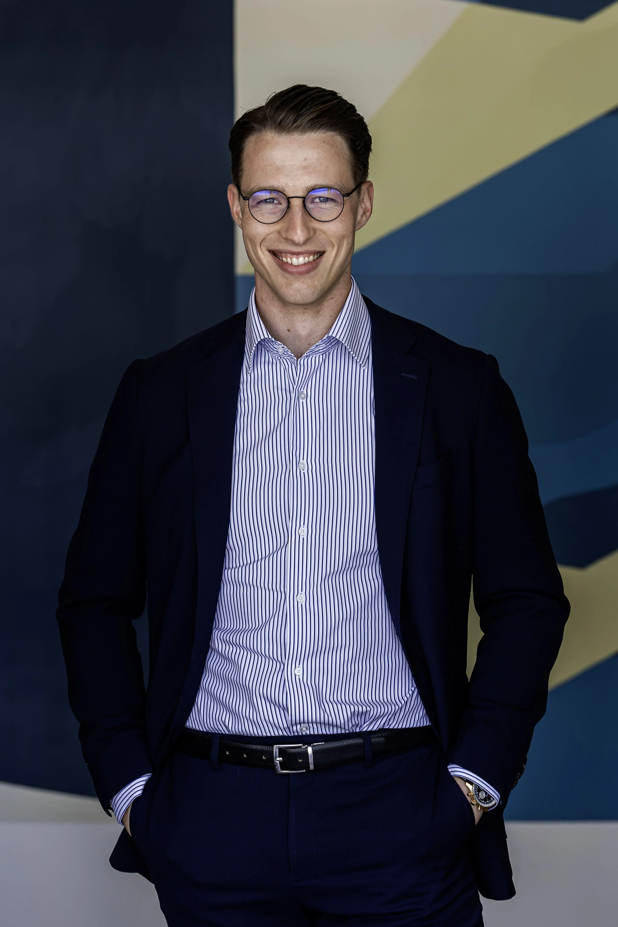 A man with brown hair, wearing glasses, a navy blue suit, and a striped shirt, smiling and standing with one hand in his pocket against a colorful geometric background.