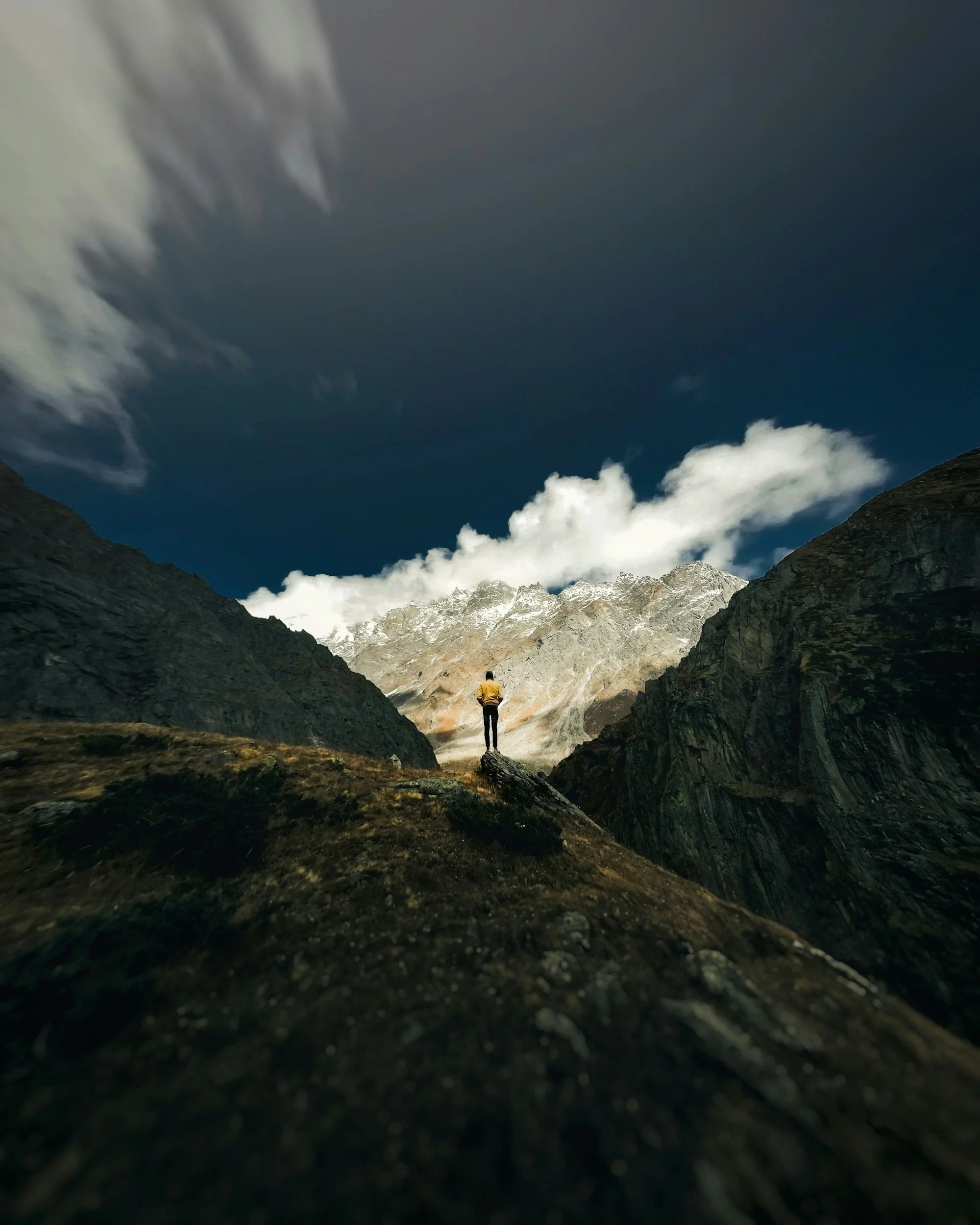A person standing on a rocky terrain in a mountainous landscape, with snow-capped peaks in the background and a cloudy sky above.