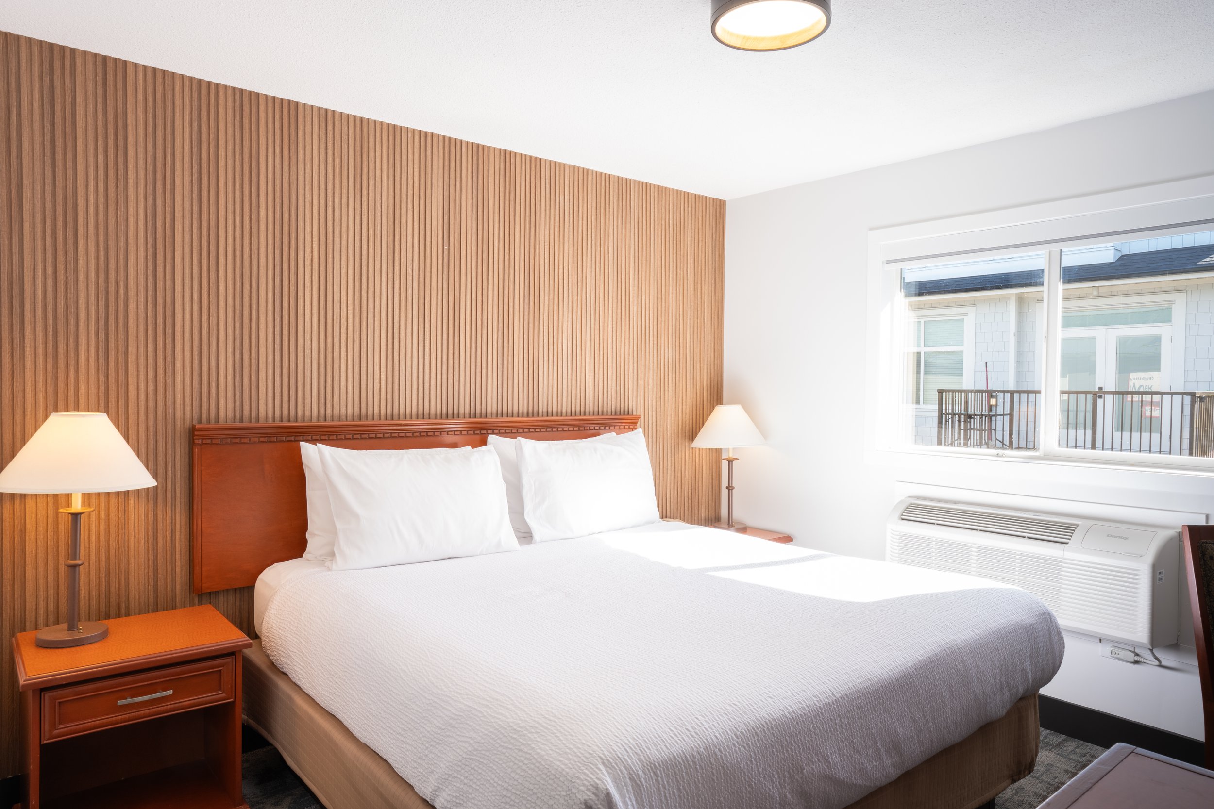 A hotel room with a bed, white bedding, and pillows. There is a wooden headboard, a nightstand with a lamp, and a large window letting in sunlight. The room has white walls and a wood-paneled accent wall behind the bed.