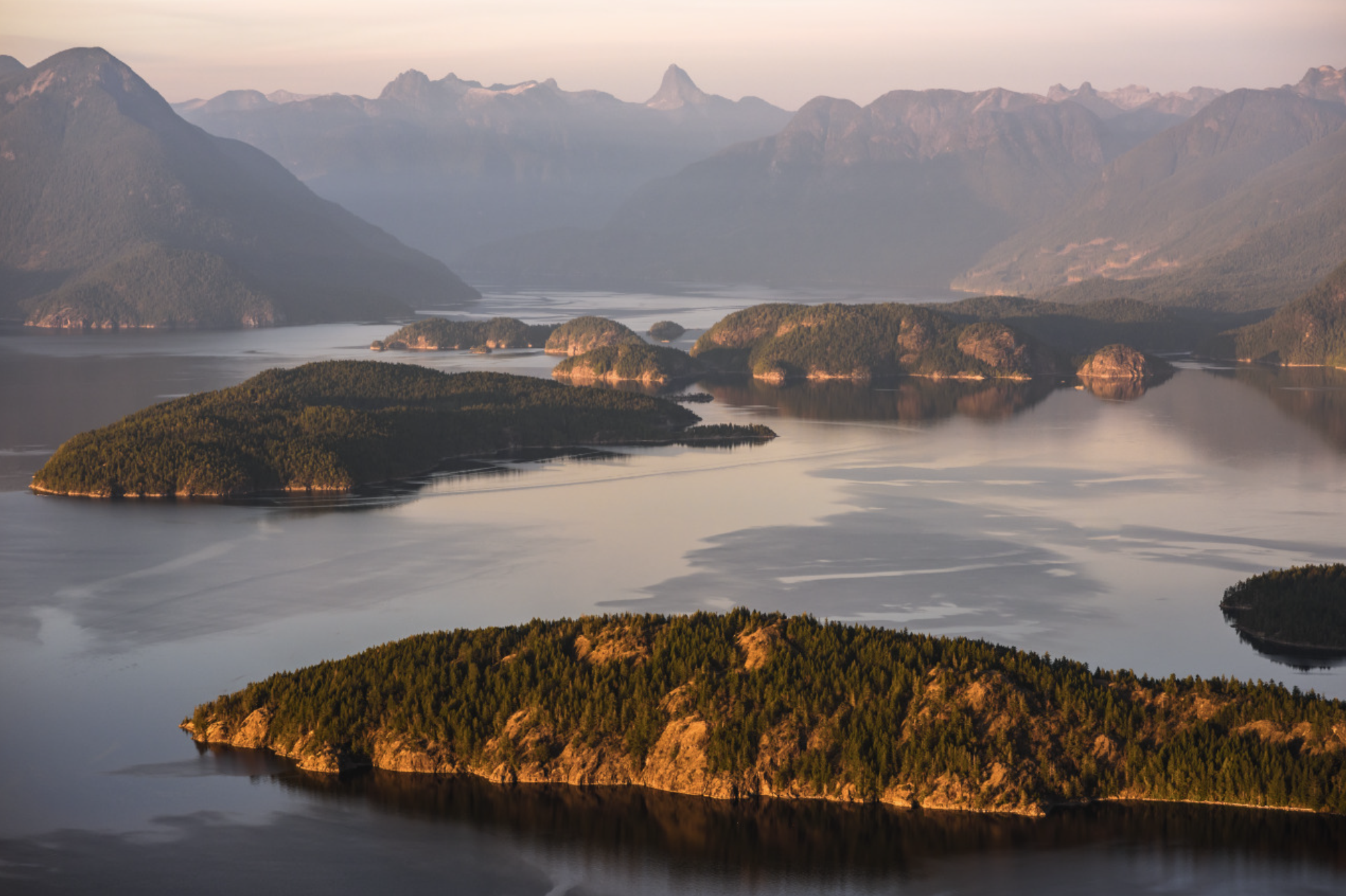 Scenic view of a mountain landscape with forests, islands, and a body of water, during a sunset or sunrise.