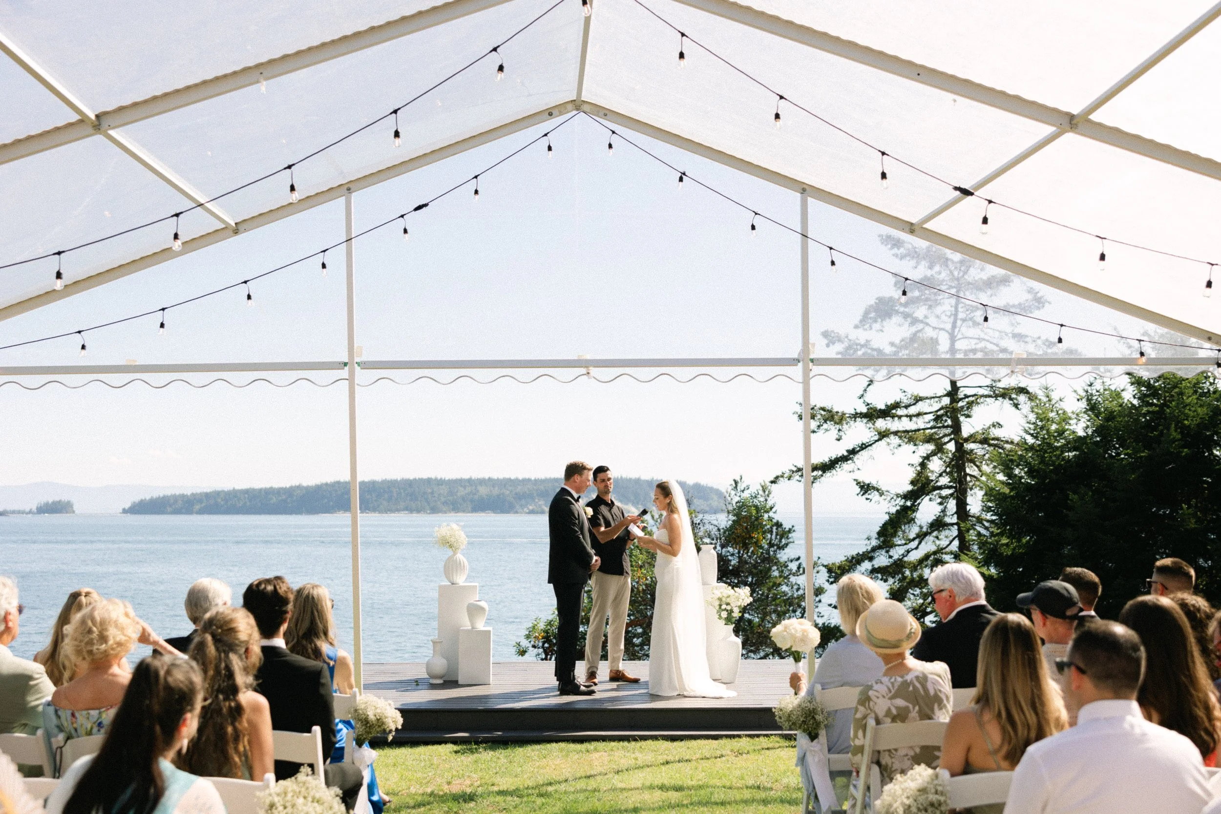 A couple gets married outdoors by a lakeside with guests seated on white chairs watching. The ceremony takes place under a white tent decorated with string lights and floral arrangements, with a scenic view of water and trees in the background.