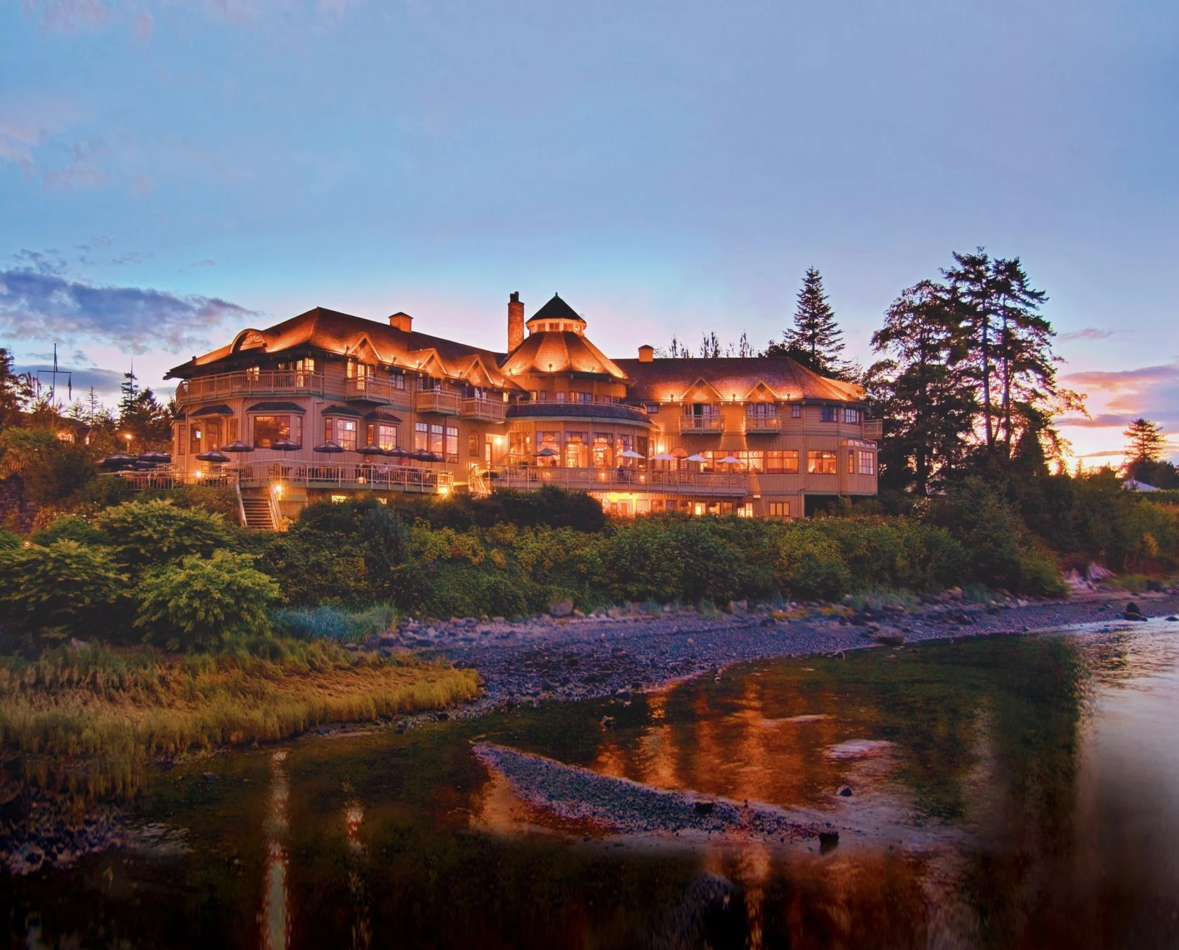 A large, multi-story house with warm lighting, surrounded by trees, on a hillside near a river at sunset.