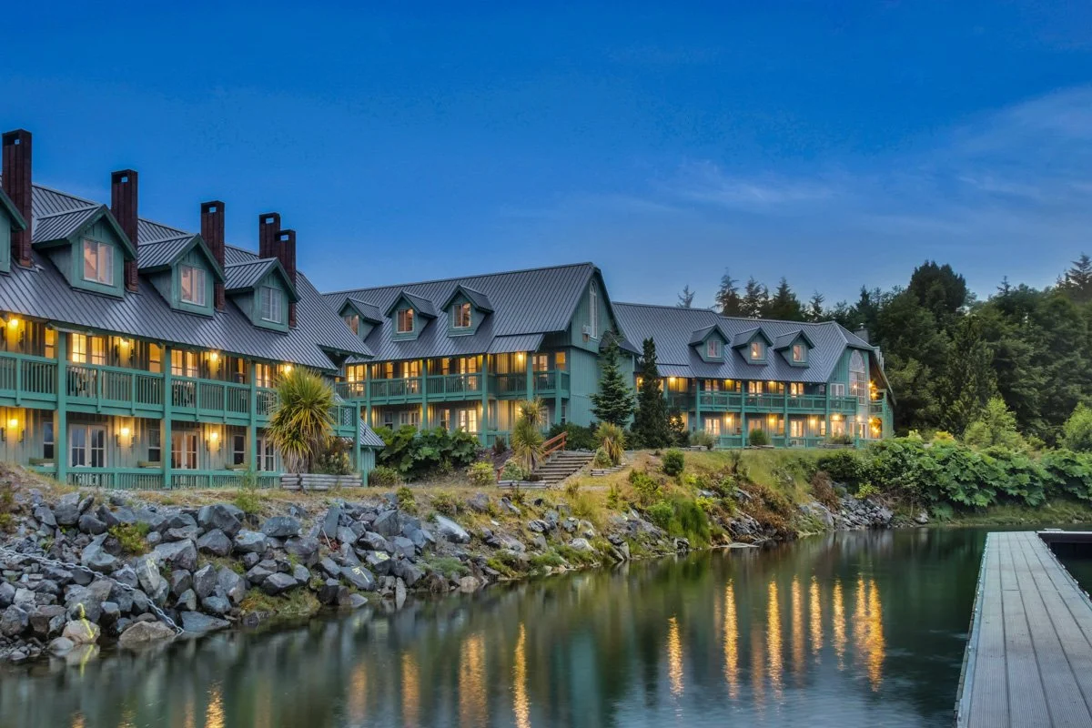 A lakeside hotel with multiple stories, teal exterior, and pitched roofs, illuminated with warm lights during twilight, surrounded by trees and rocks.