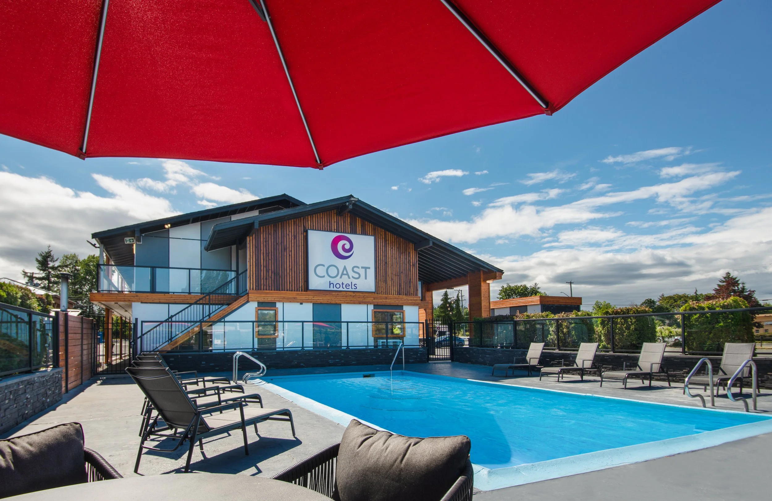 Outdoor pool area at Coast Hotels with lounge chairs, a building with a sign, and a large red umbrella in the foreground.