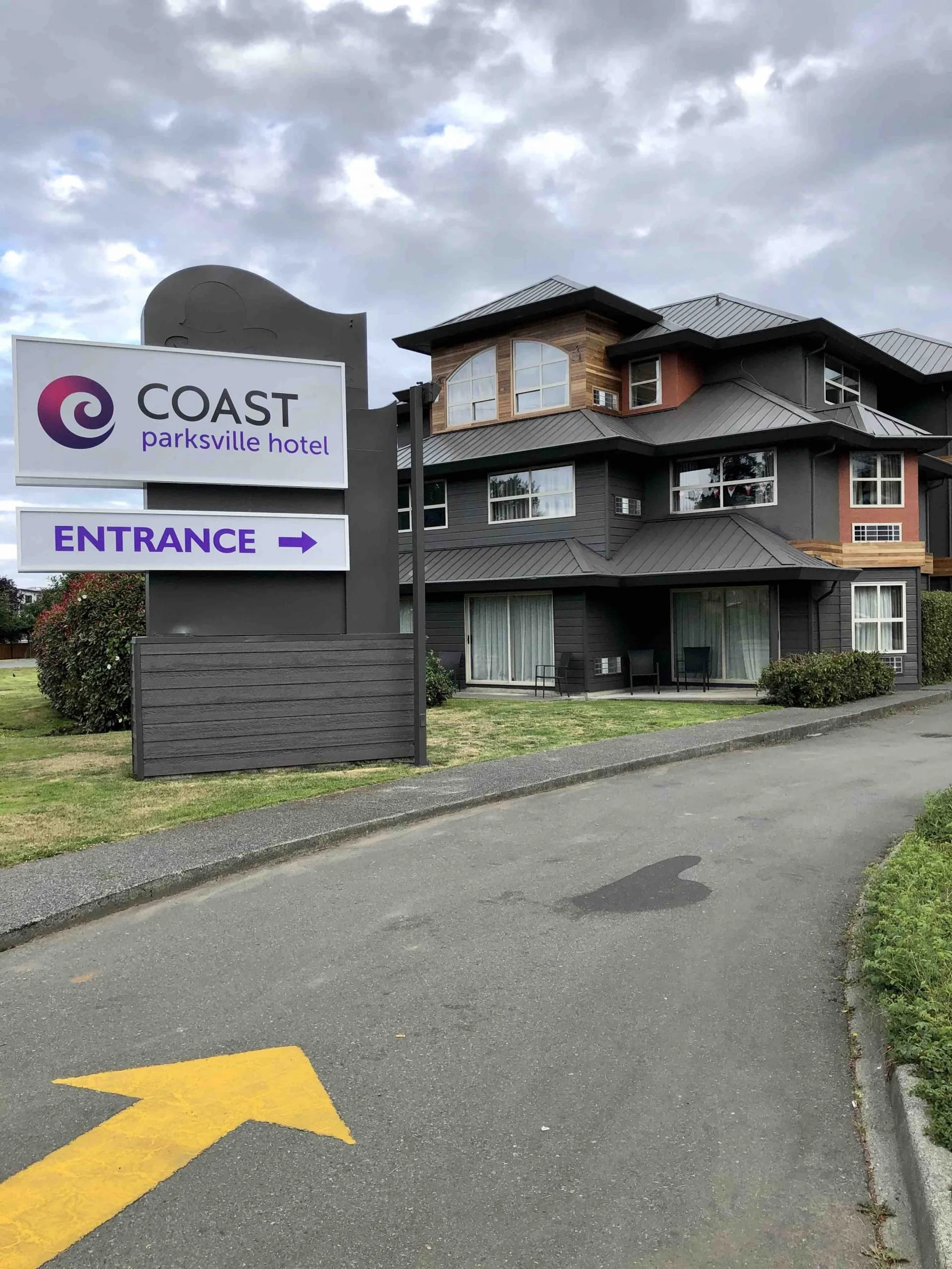 A parking lot with a yellow arrow painted on the asphalt pointing left, next to a curb with grass and bushes. In the background, there is a multi-story building with dark gray and wood siding, multiple windows, and a prominent sign that reads 'COAST parksville hotel' with an arrow indicating the entrance.