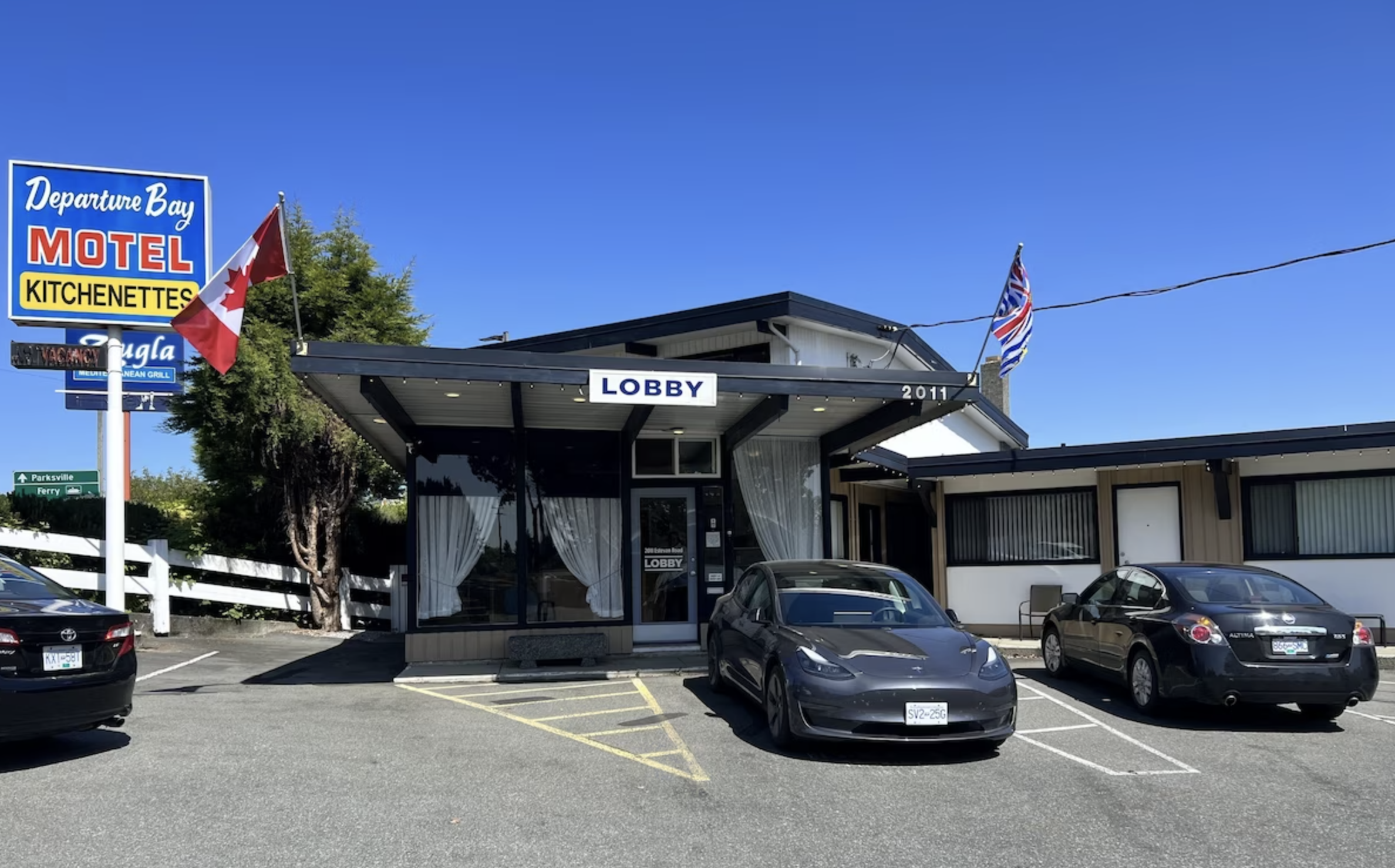 Front view of a motel named 'Departure Bay Motel' with parking spaces in front, surrounded by cars, flags, and a clear blue sky.