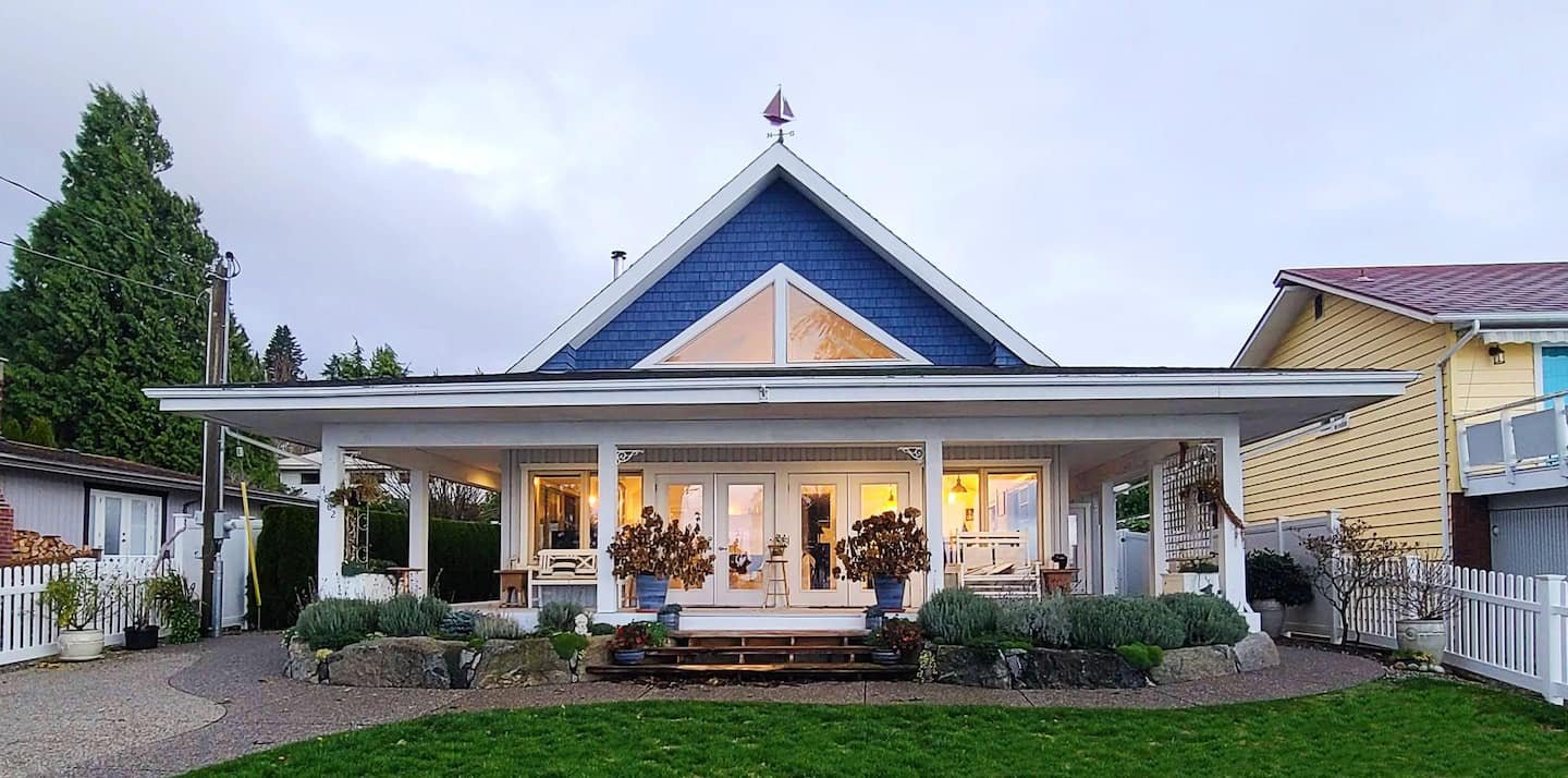 Front view of a house with a porch, white fences, and a garden, during dusk with lights on inside.
