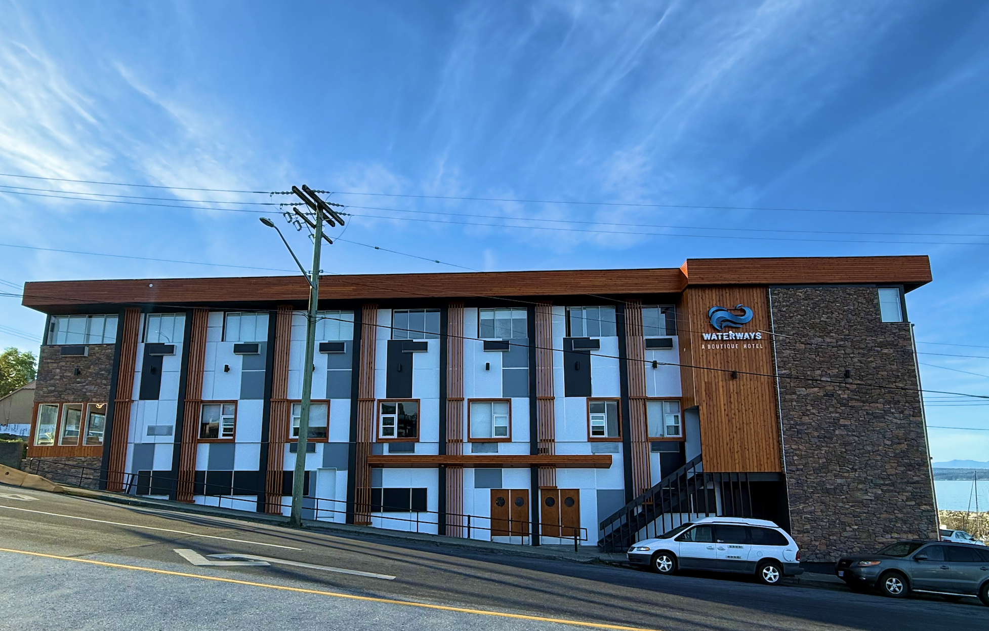 A modern hotel building named Waterways Boutique Hotel with a unique exterior design featuring horizontal wooden slats and large windows, located along a street with parked cars and utility poles under a clear blue sky.