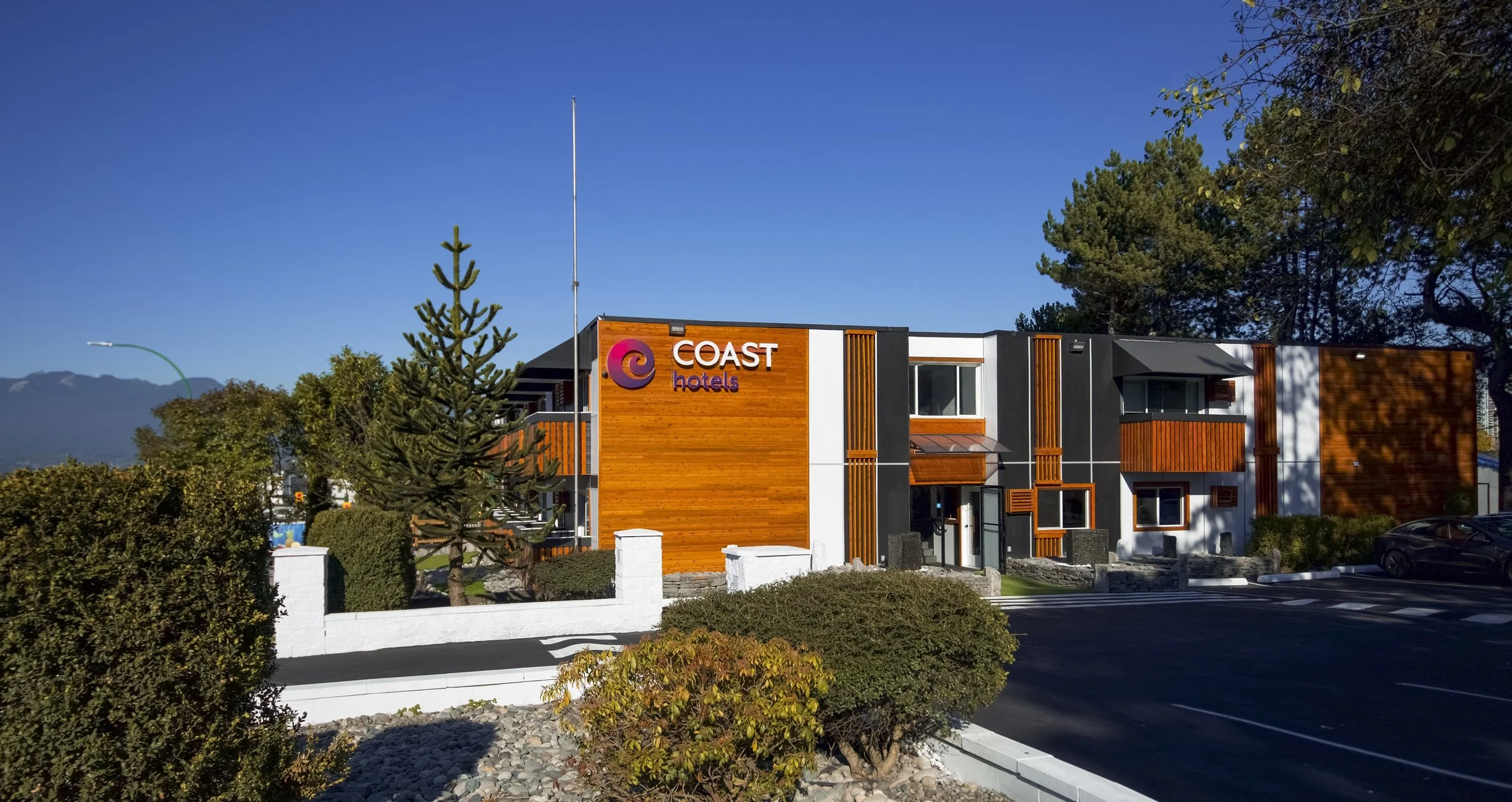 Exterior of Coast Hotels building with modern architecture, wooden accents, and signage, surrounded by trees and greenery under a clear blue sky.