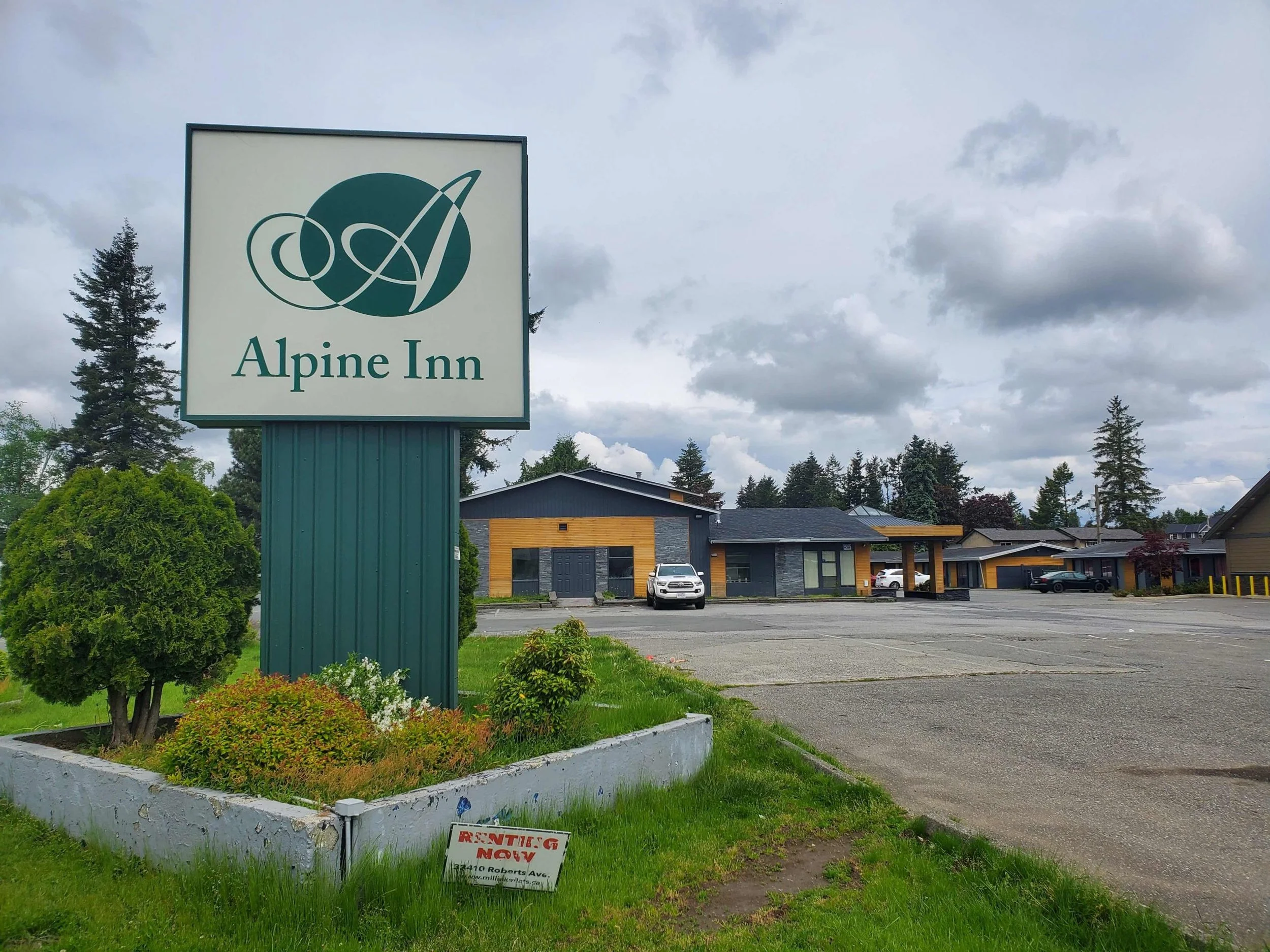 Sign for Alpine Inn in a parking lot with nearby residential buildings, trees, and cloudy sky.