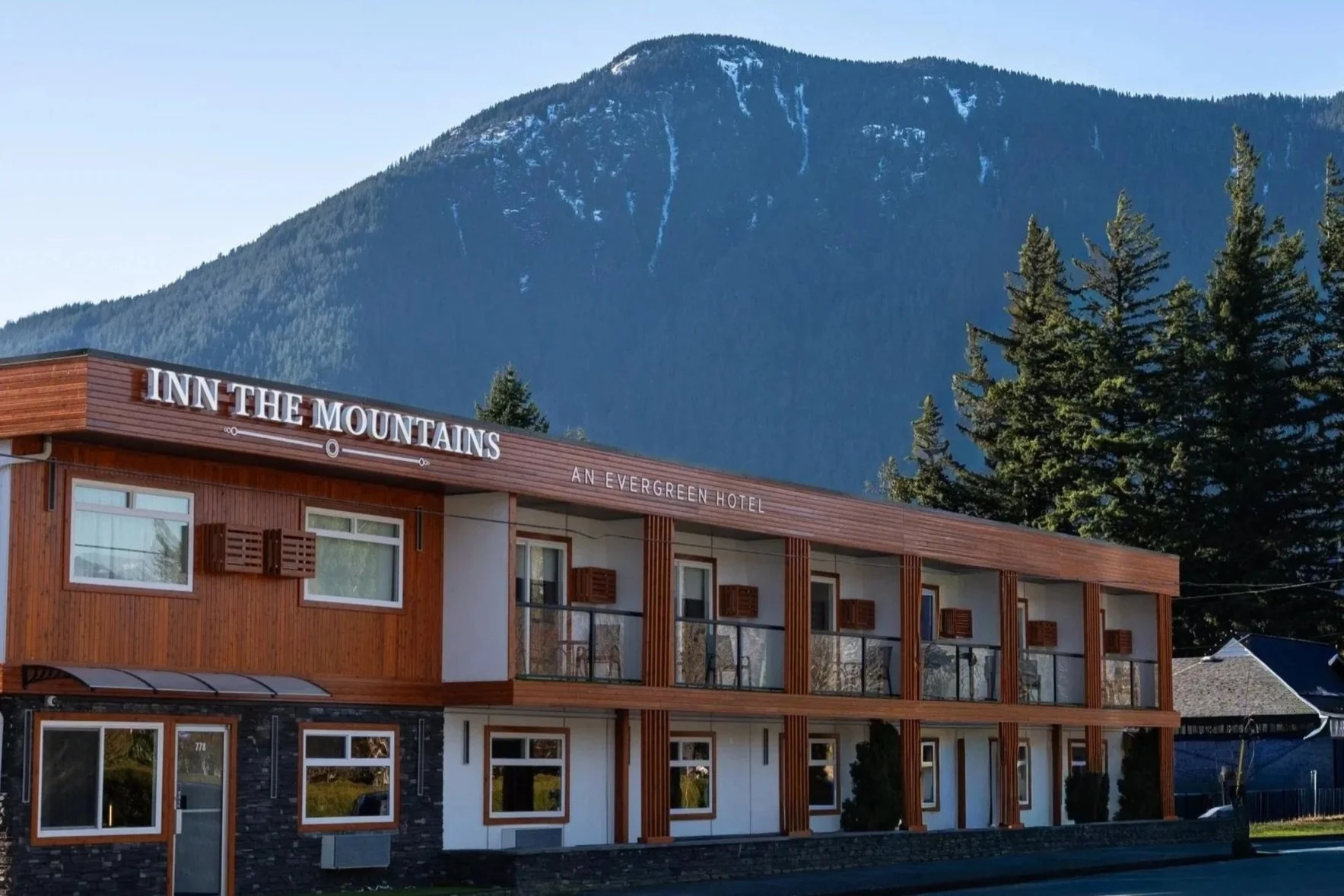 A modern, multi-story hotel called Inn the Mountains with the subtitle 'An Evergreen Hotel', set against a backdrop of a large mountain with patches of snow and dense green trees.