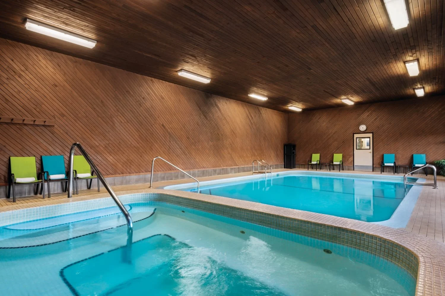 Indoor swimming pool with two sections, surrounded by a wooden wall and chairs along the side, illuminated by ceiling lights.