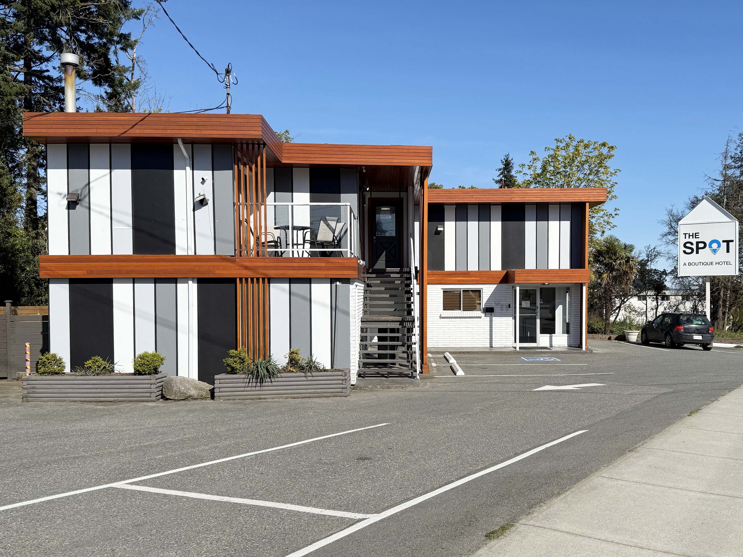 A modern, multi-story building with a black and white exterior, wooden accents, and a balcony with chairs. The parking lot in front is mostly empty, with a few cars, and a sign reads "The Spot A Boutique Hotel." There are trees and a clear blue sky in the background.