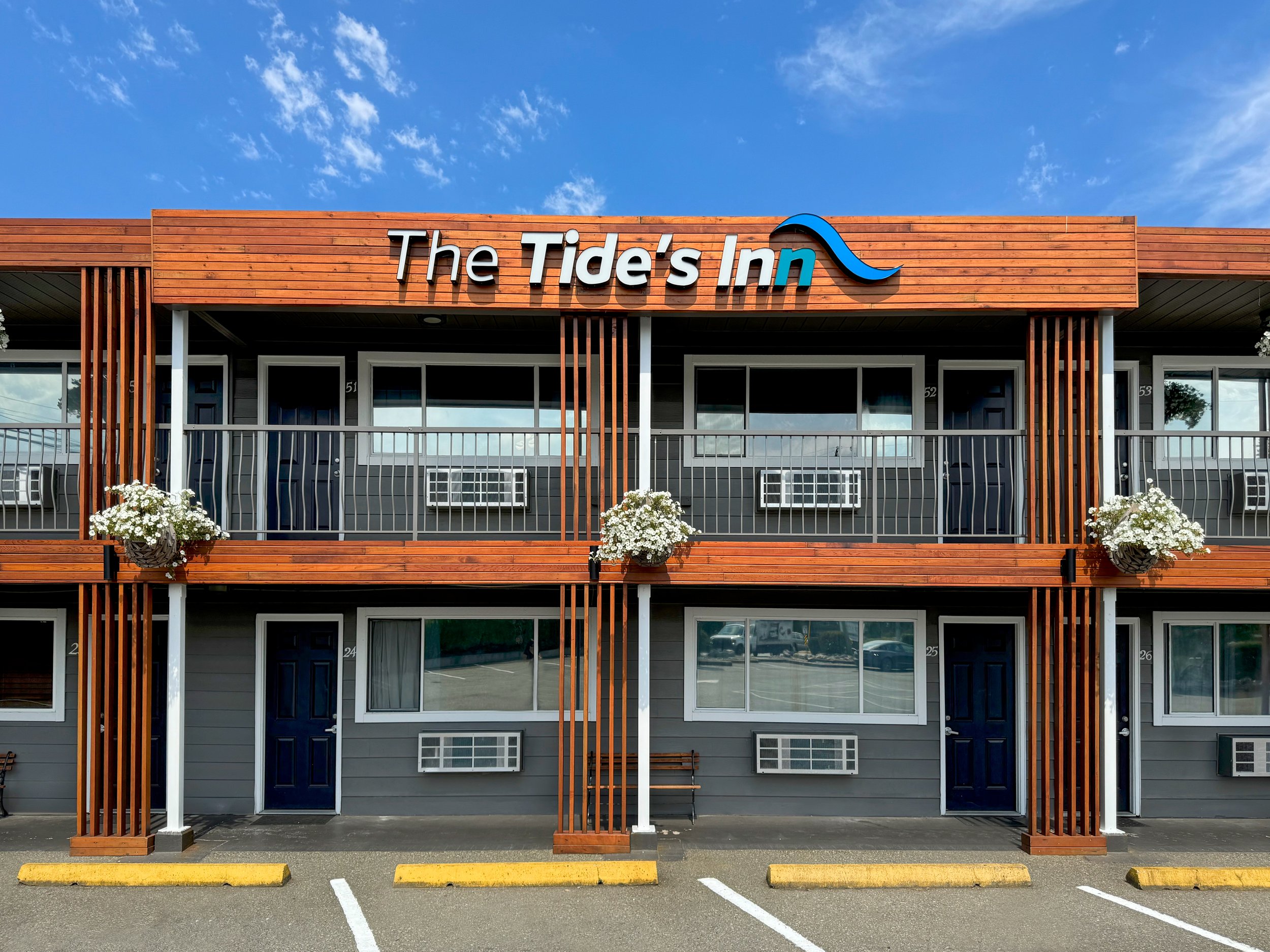 Front view of a two-story motel building with a sign reading "The Tide's Inn" featuring a blue wave design, gray walls, black doors, white-framed windows, and hanging flower baskets, under a blue sky with scattered clouds.
