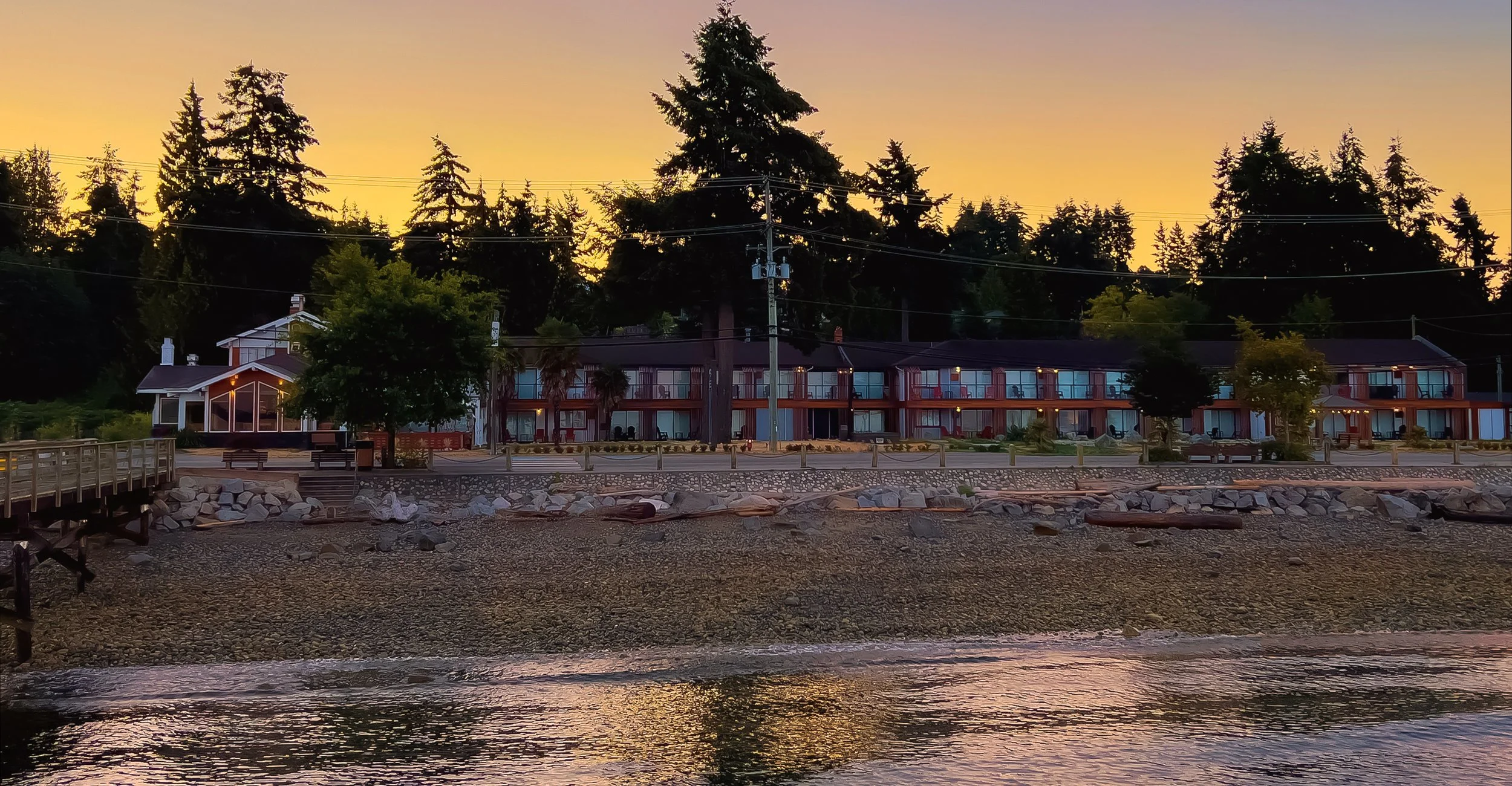 Scenic view of waterfront apartments during sunset with trees, utility poles, and rocks along the shoreline.
