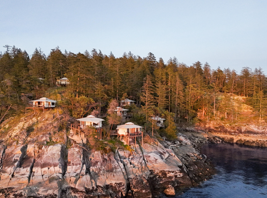 Several small vacation cabins built on rocky cliffs overlooking the ocean, surrounded by trees at sunset.