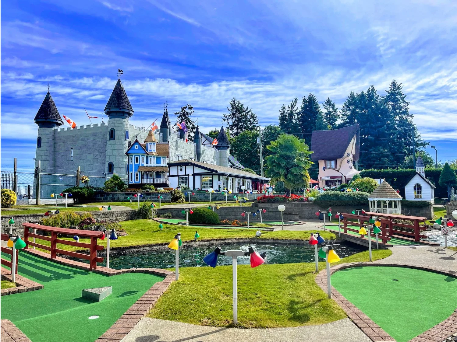 Miniature golf course with a castle-themed building in the background, colorful flags, a pond, a small bridge, and well-maintained landscaped grounds under a partly cloudy sky.
