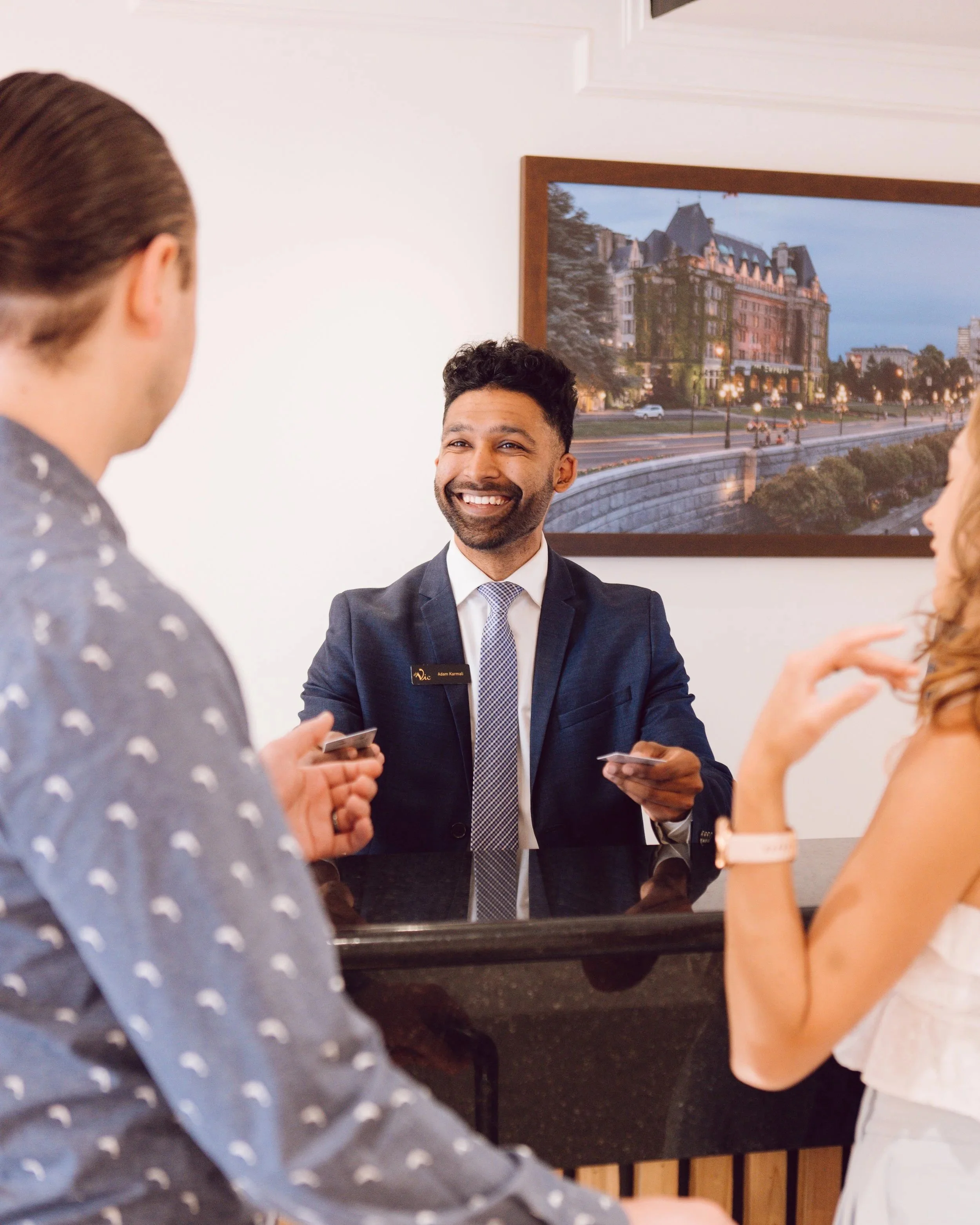 A man in a suit and tie smiling and sitting at a black table with two women, one wearing a watch and the other wearing a white shirt, hands visible. There is a framed picture of a cityscape with buildings and a river in the background. The scene appears to be a professional or social setting.