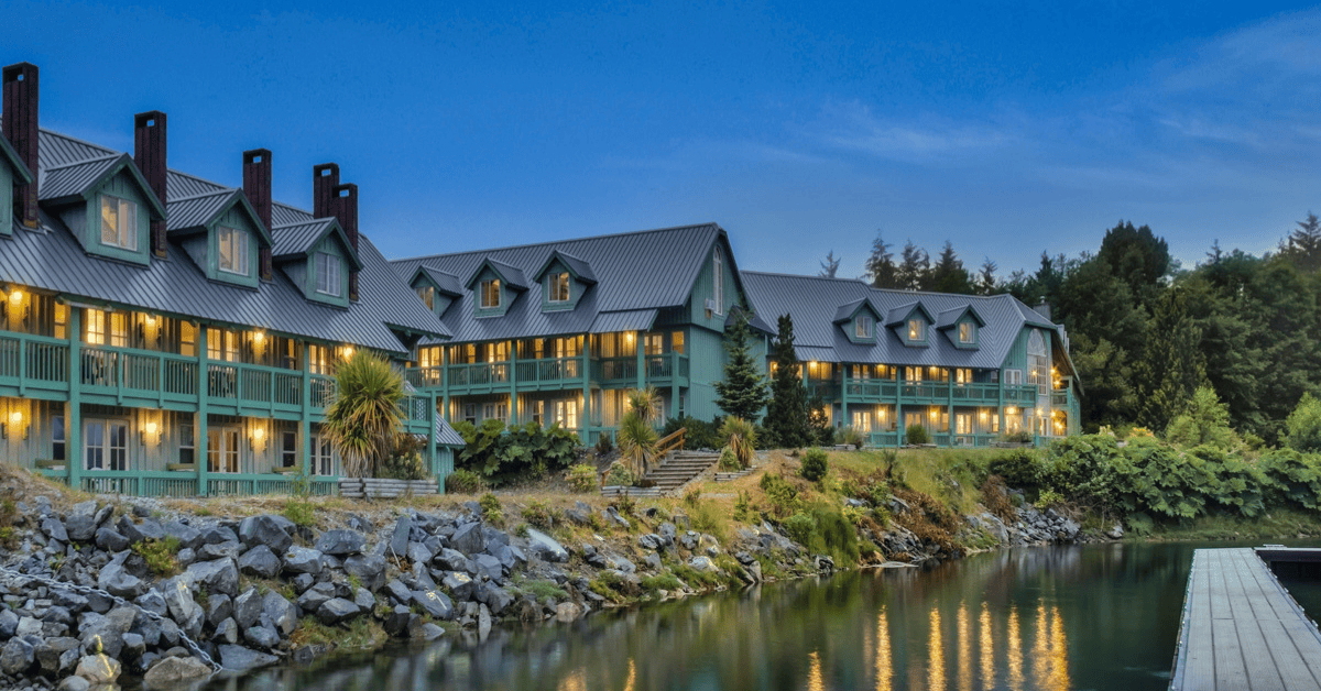 A large, multi-story hotel or lodge with a green exterior and multiple peaked roofs, situated on a rocky riverbank with a dock, surrounded by trees, dusk or early evening, with lights on inside.