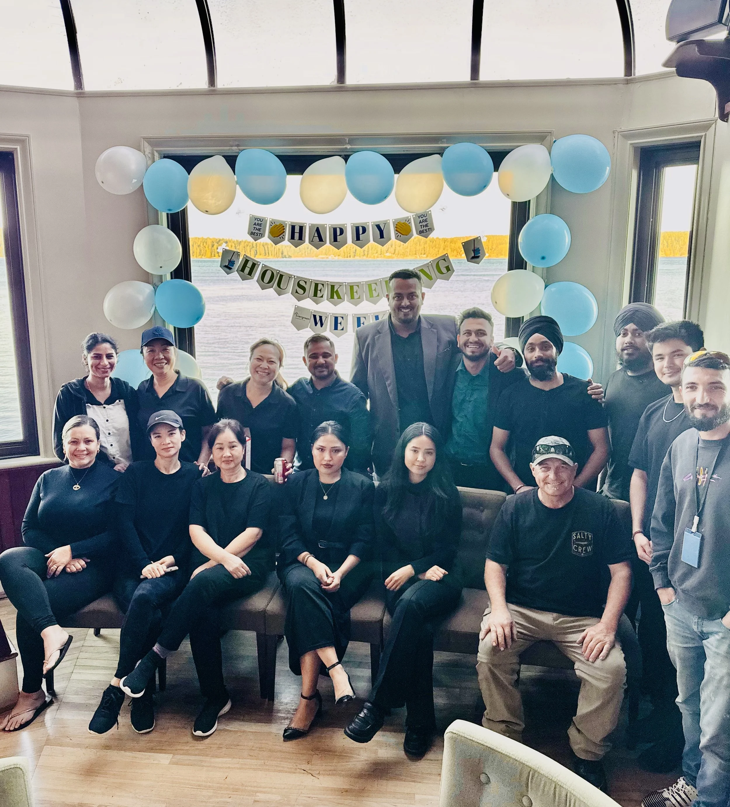 A group of diverse people smiling and posing indoors at a celebration. Decorations include blue and yellow balloons and a banner that reads 'HAPPY HOUSEKEEPING WEEK'. Large windows reveal a body of water and trees outside.