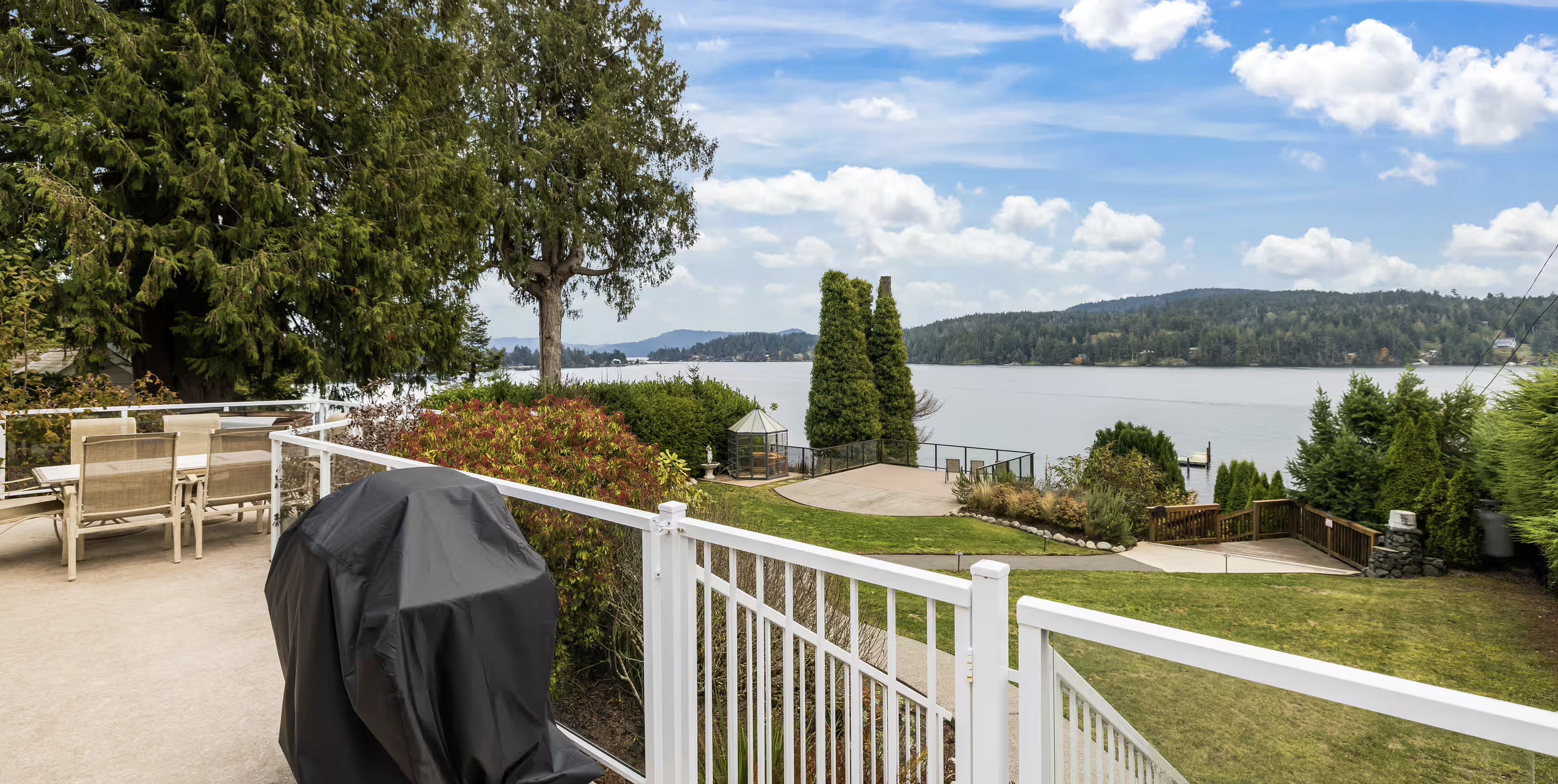 View from a backyard patio overlooking a lake with trees and hills in the distance, cloudy sky, outdoor dining furniture, and a barbecue grill.