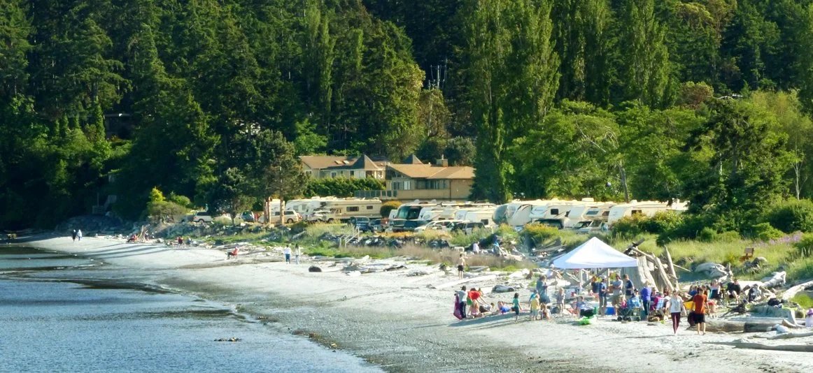 People gathered on a beach near the shoreline with trees and houses in the background, some under a white canopy tent.