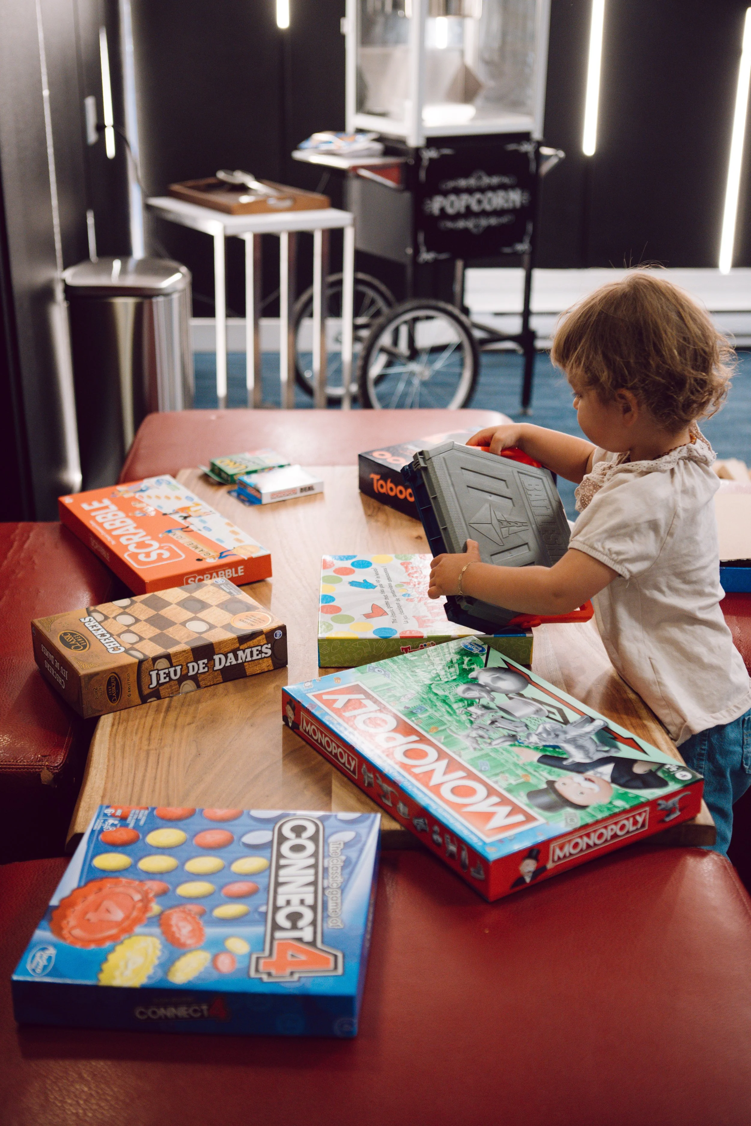A young girl with light brown hair playing a tabletop board game in a cozy setting. Several board games are visible on the table including Monopoly, Connect 4, Scrabble, and a puzzle game. In the background, there is a popcorn cart and a wheelchair.