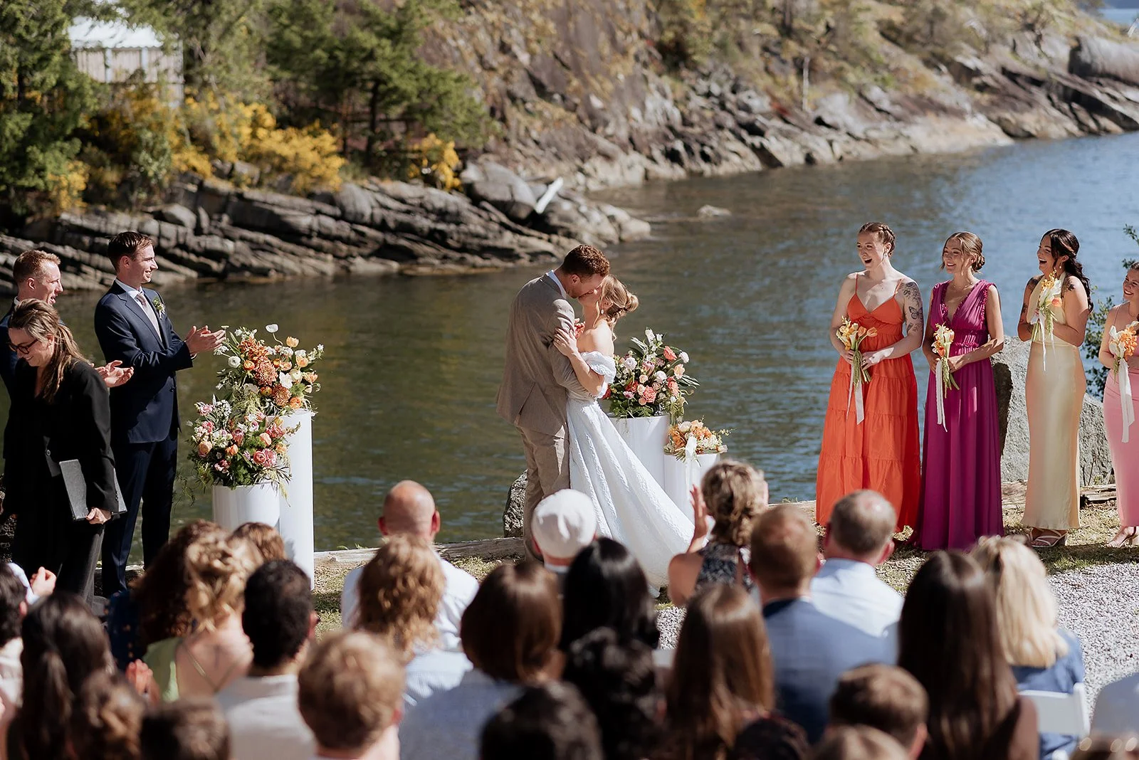 A wedding ceremony by a river with a groom and bride kissing, surrounded by bridesmaids and groomsmen, with an audience watching on.