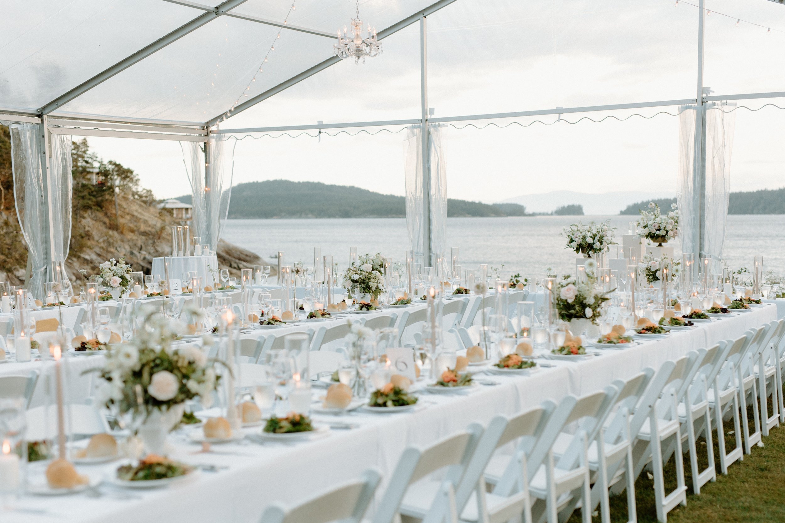 Long banquet table set with white tablecloths, floral centerpieces, candles, and place settings, under a transparent tent overlooking a body of water and distant hills.