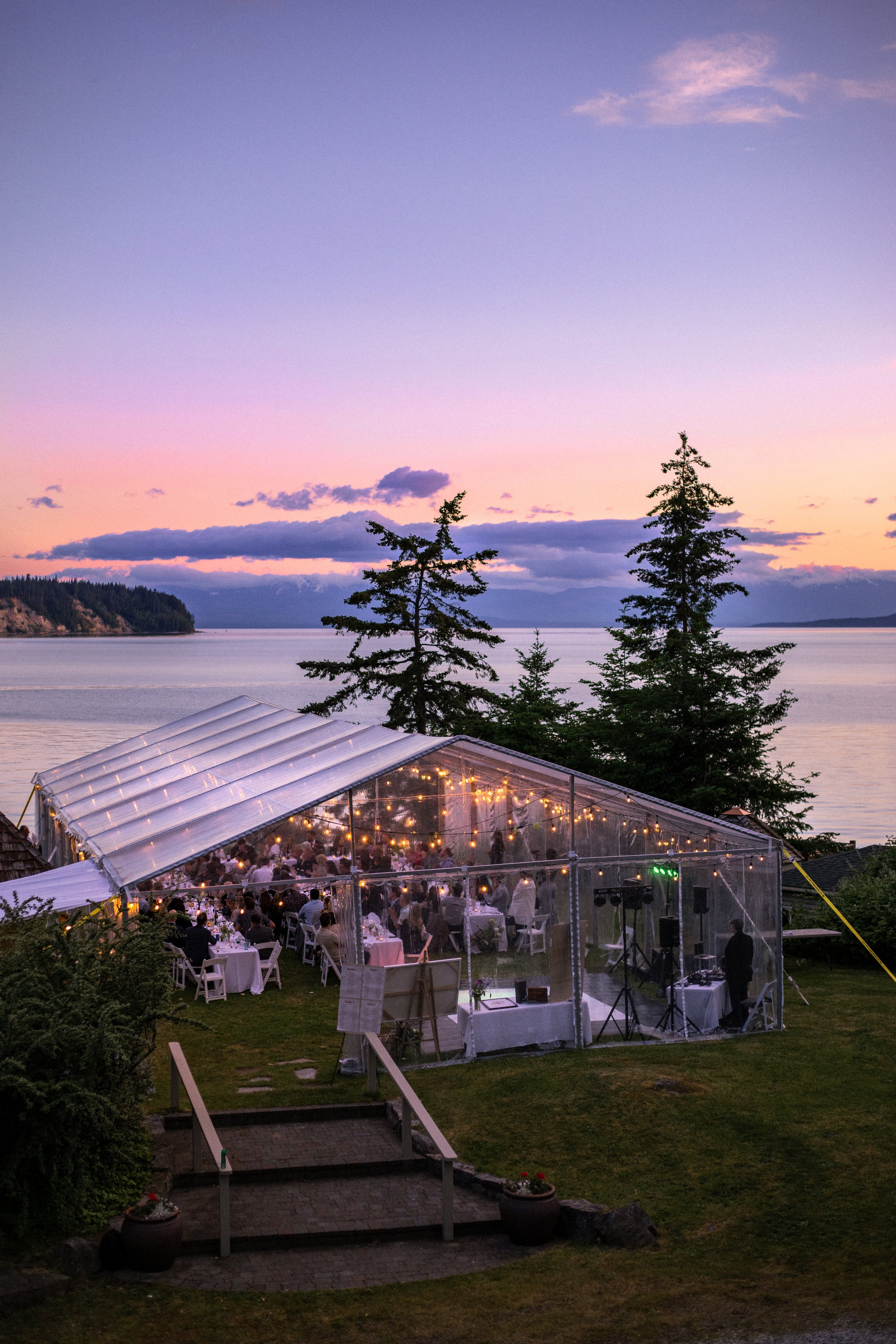 Outdoor event tent with string lights during sunset, overlooking a lake with trees and a partly cloudy sky.