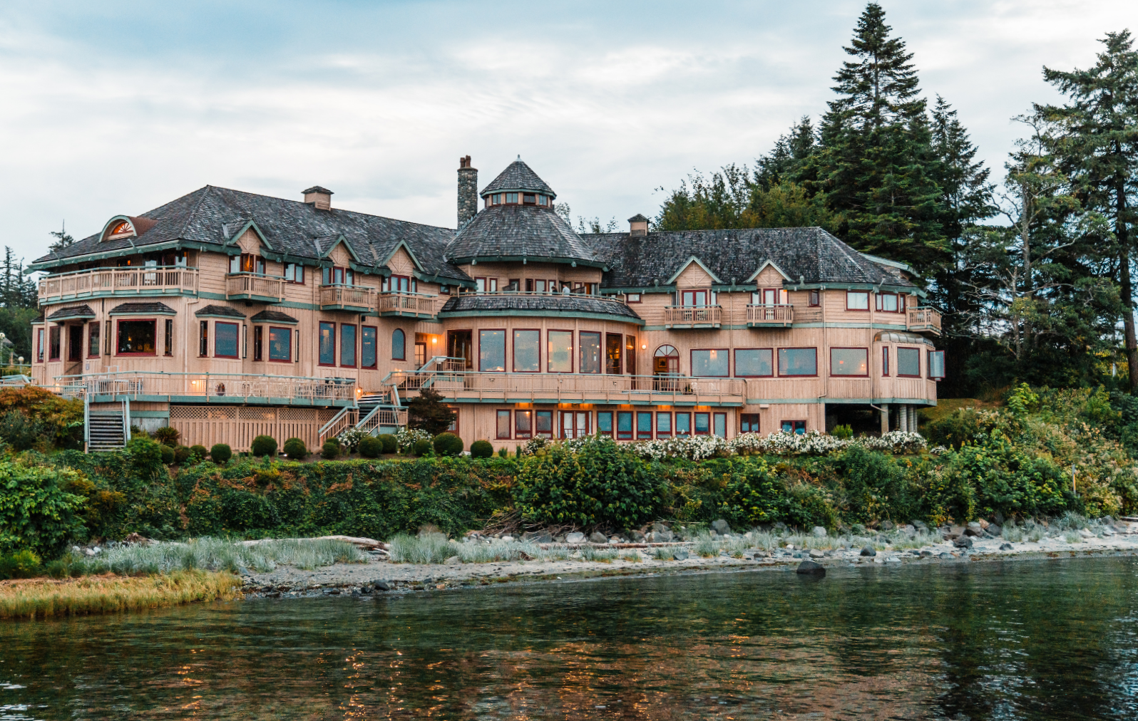 A large, multi-story house with numerous windows, balconies, and a turret, situated by a body of water and surrounded by trees and shrubs.