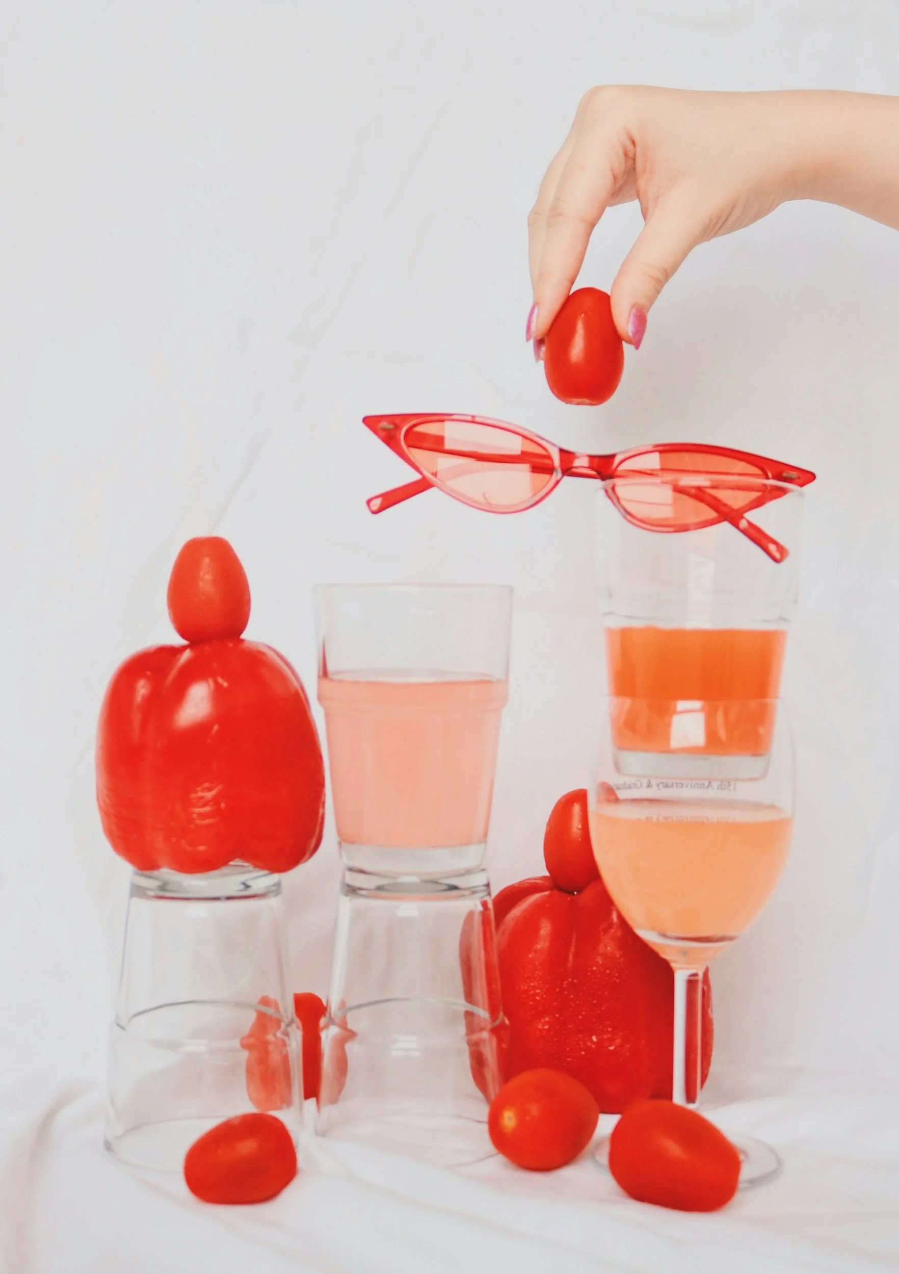 Various glassware filled with a pink drink, with cherry tomatoes and a red bell pepper arranged around and on top of the glasses, and a hand holding a cherry tomato above the glasses.