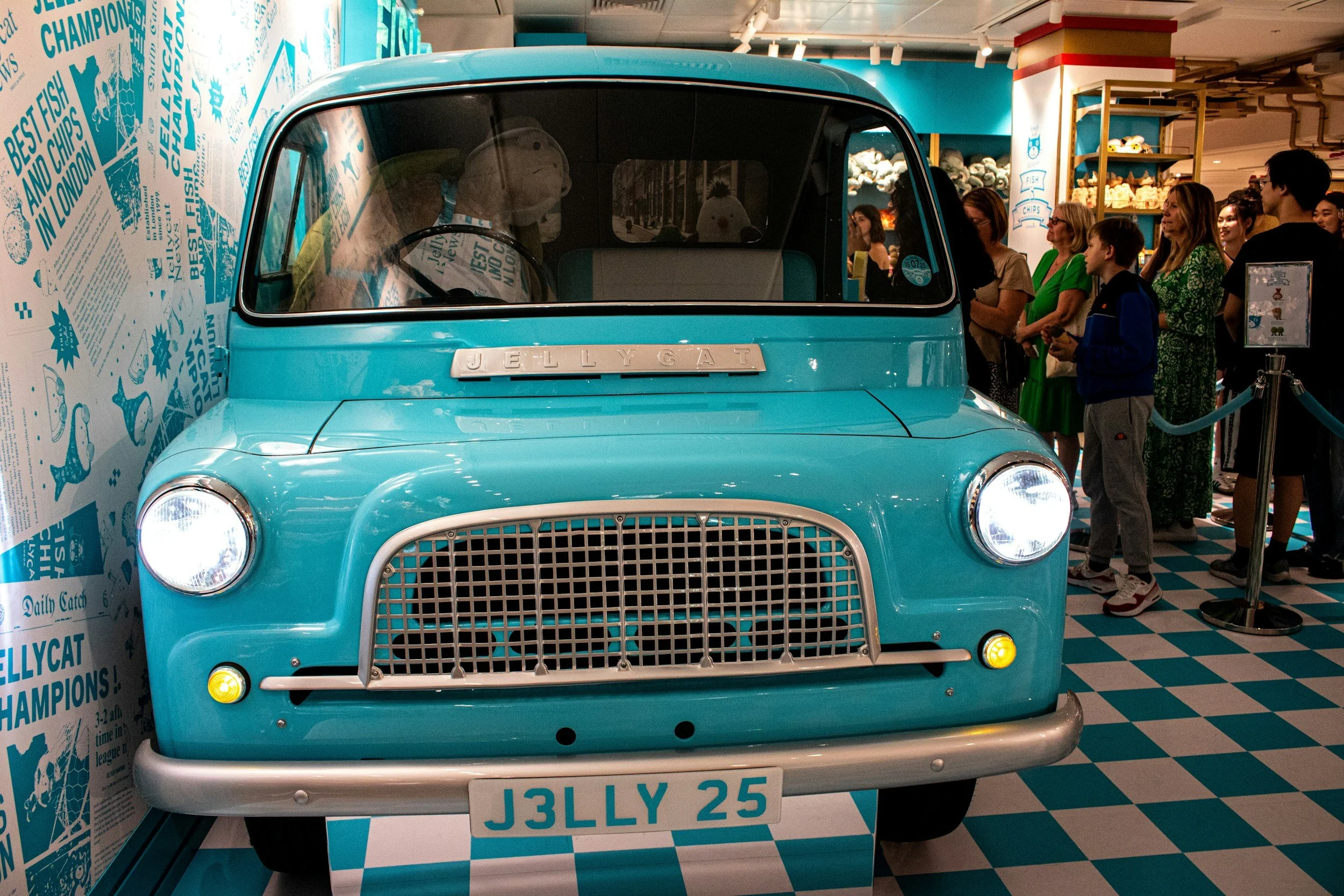 A turquoise vintage-style car is displayed indoors at a public event. The car is part of a themed exhibit, with a crowd of people, including children and adults, standing in line behind a barrier to view it. The background features a wall with blue and white graphics and text.