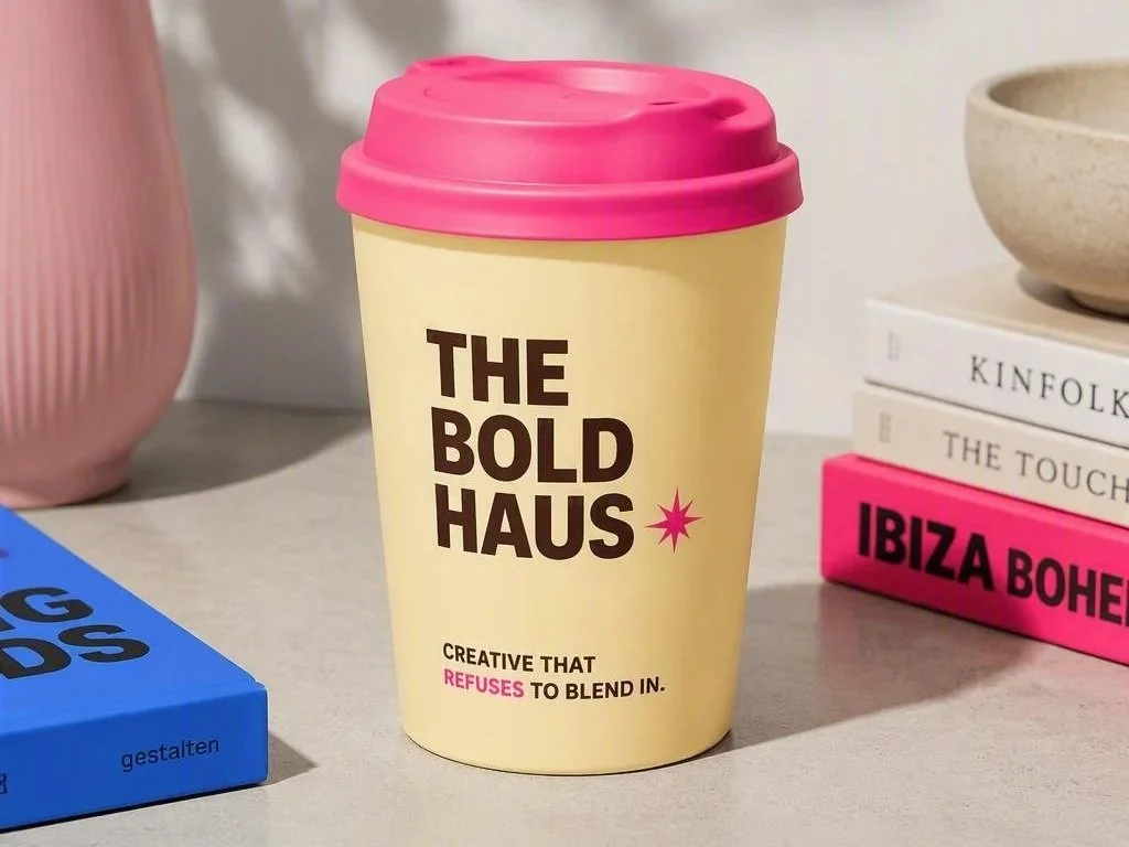 A beige disposable coffee cup with a pink lid and bold black text that reads 'The Bold Haus' and a pink star. Below, it says 'Creative that refuses to blend in.' The cup is on a table with a pink vase, a white book, and a pink book in the background.