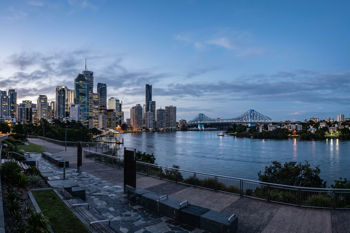 View of a city skyline with tall buildings along a river at dusk, with a bridge in the distance and a waterfront park in the foreground.