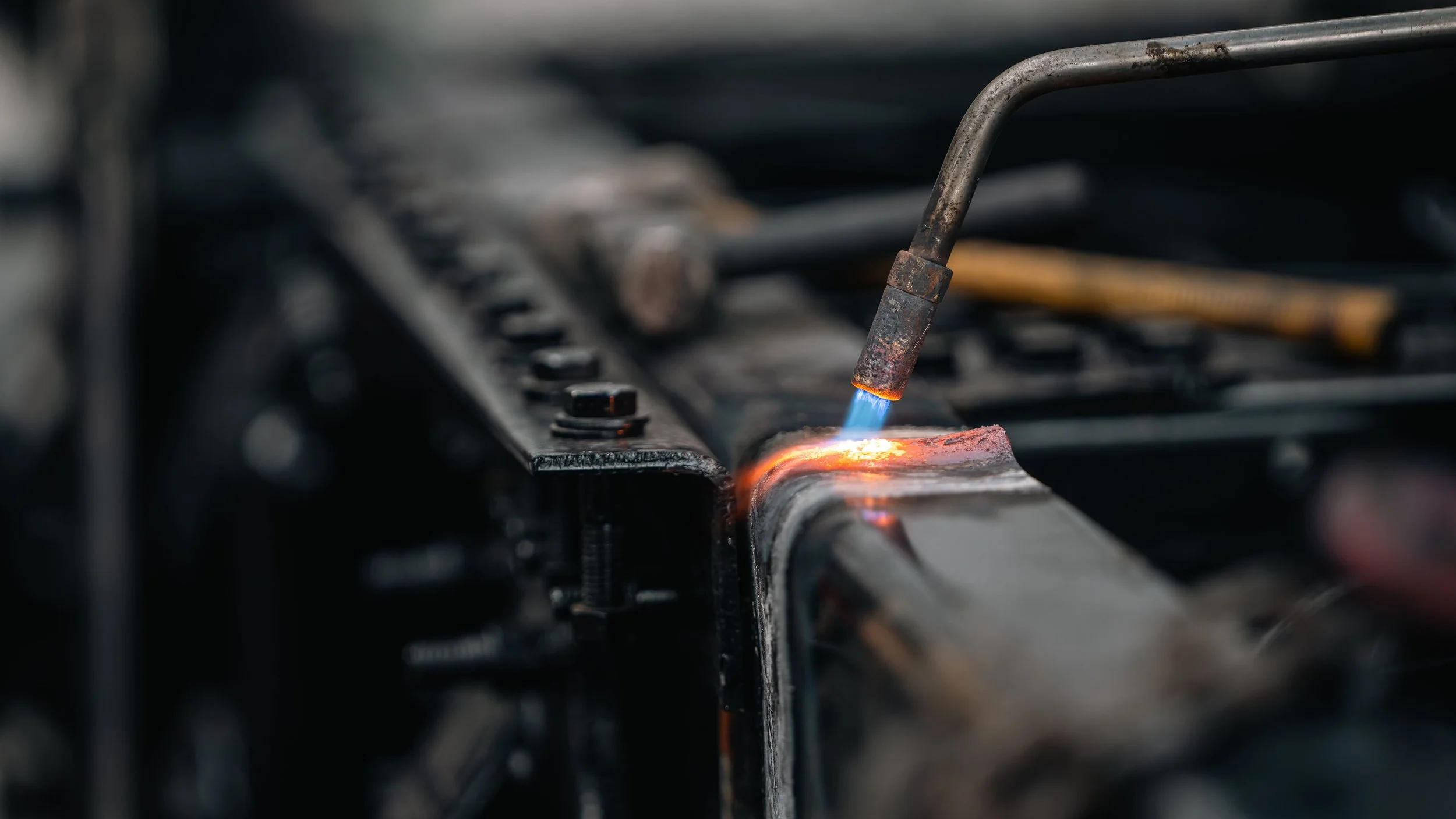 A welding torch is fusing metal parts on a dark industrial workbench, with bright blue and orange flames illuminating the scene.