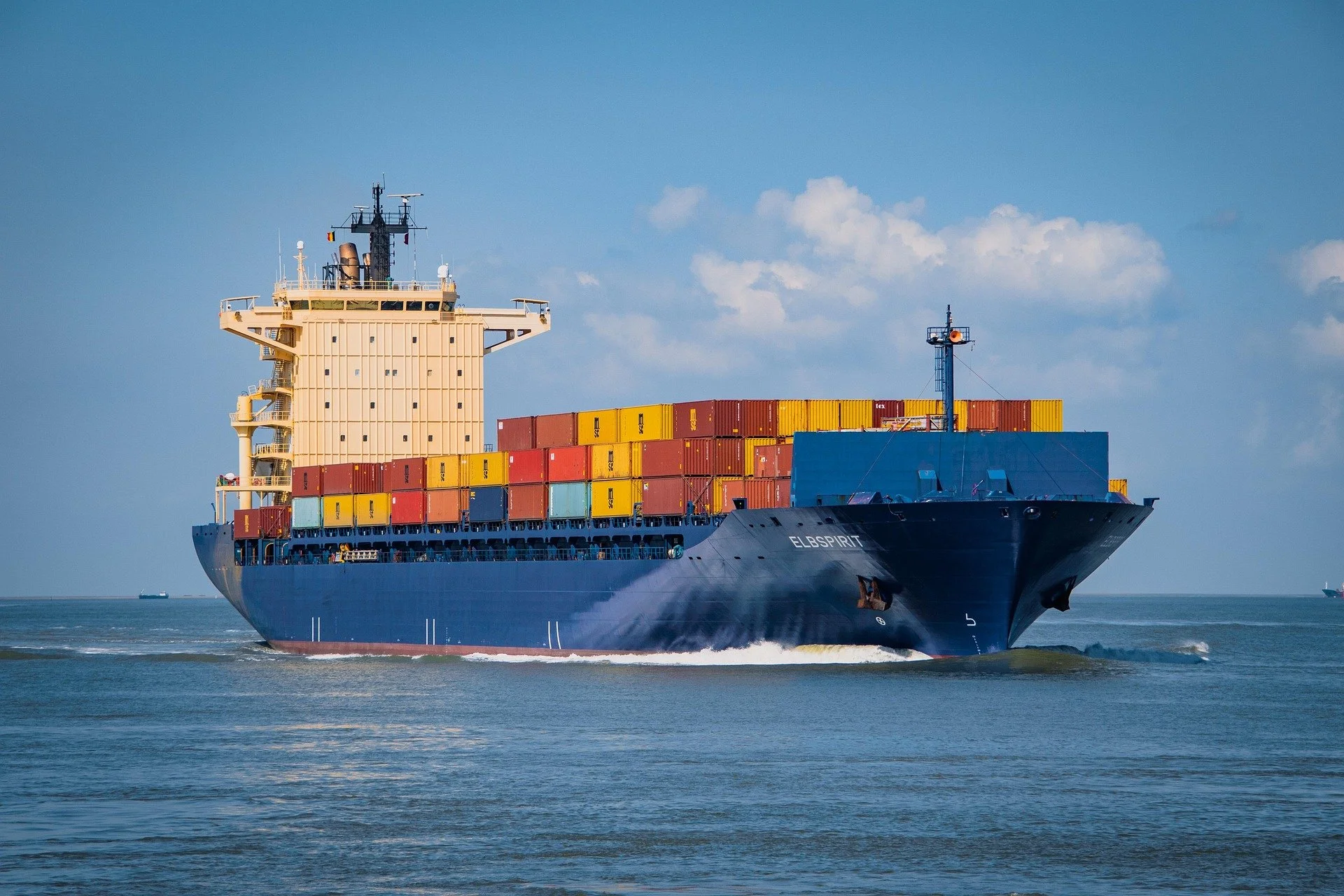A large cargo ship with colorful containers sailing on the ocean with a cloudy sky in the background.