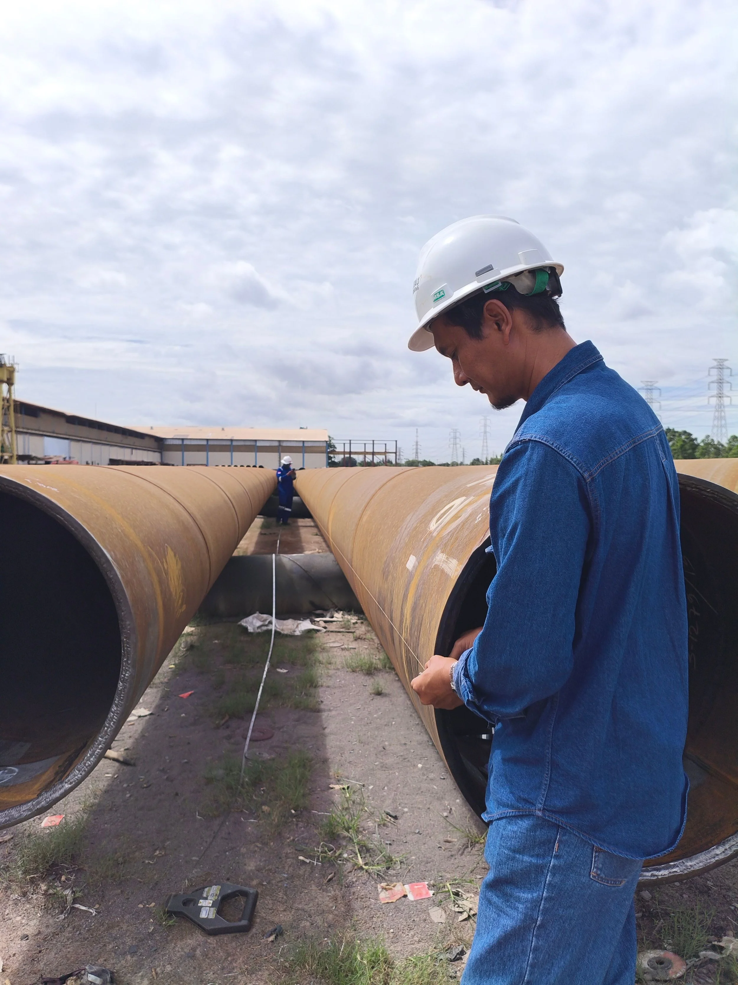 A worker in a denim jacket and white safety helmet stands next to large underground pipelines at an industrial site, with cloudy sky and power lines in the background.